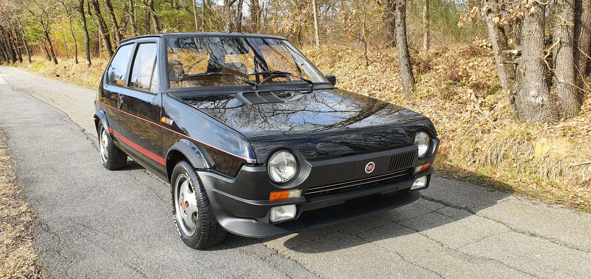 Fiat Ritmo 桌布 壁紙 — 一輛亮黑色嘅快意 Uno Turbo 停泊喺一條裂開嘅瀝青路上，兩旁係秋天嘅禿樹。