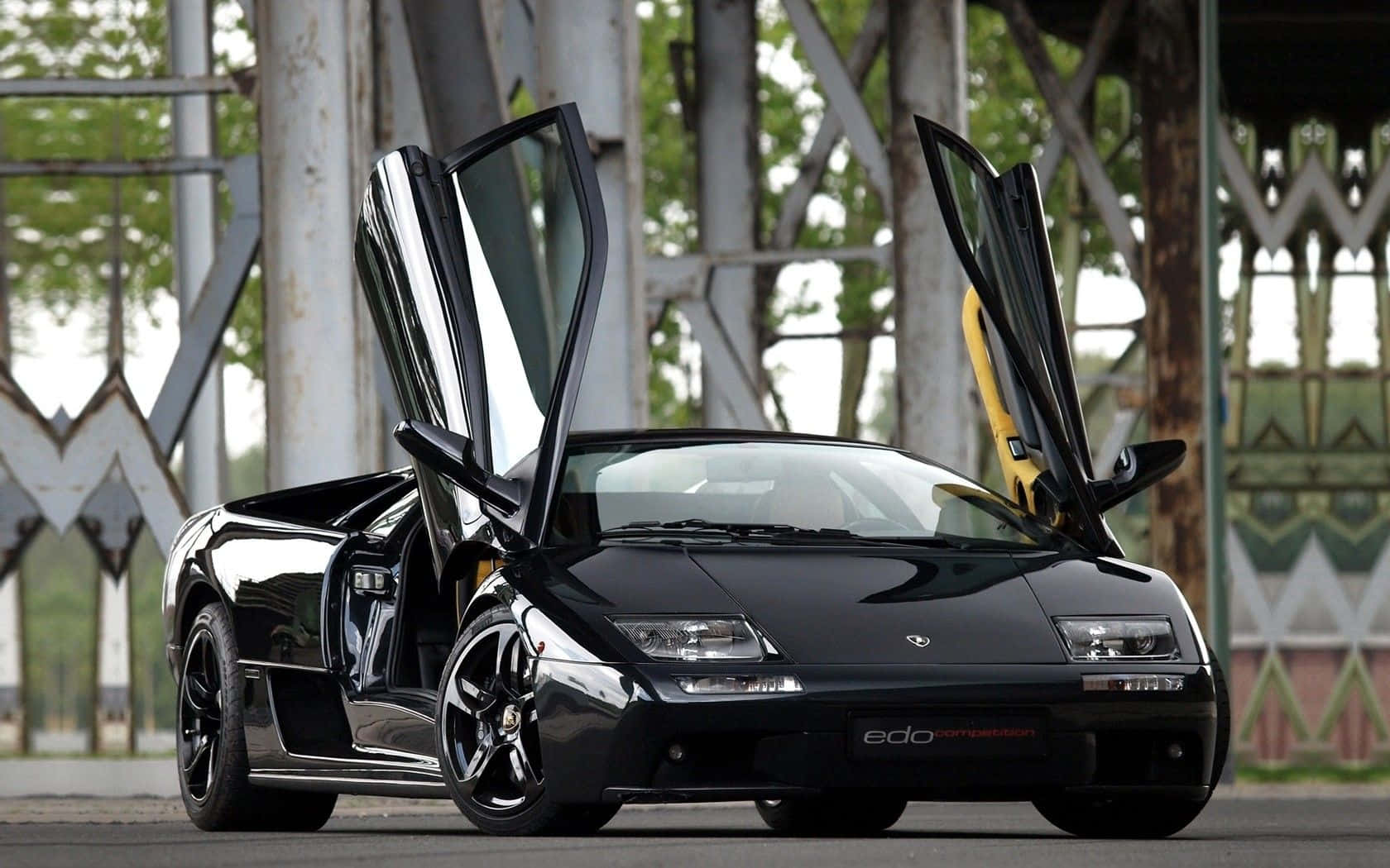 Lamborghini Diablo 桌布 壁紙 — 一架亮黑色林寶堅尼 Diablo 打開鷗翼車門，停喺金屬橋下面，背景有綠色植物。