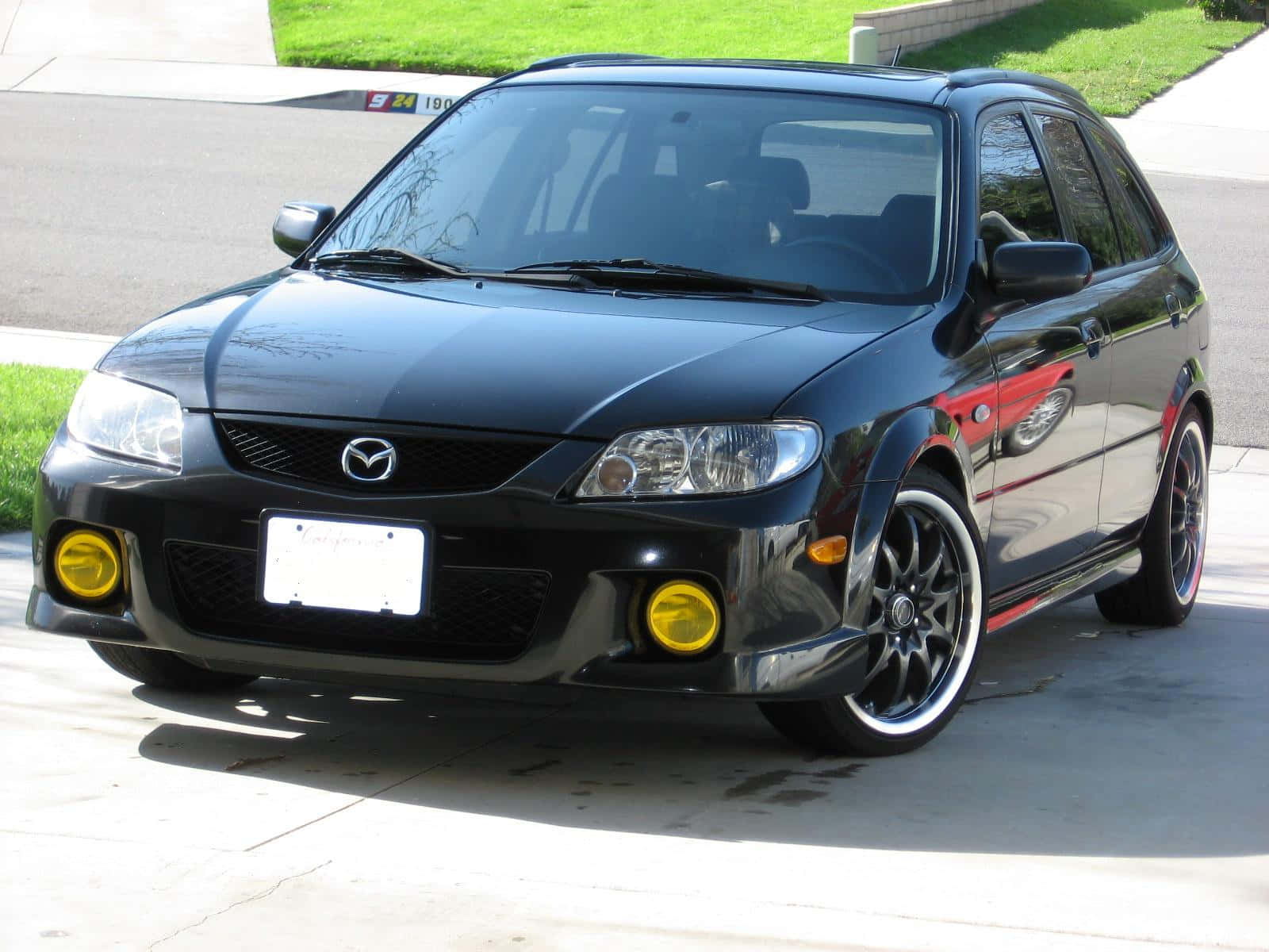 Mazda Protege 桌布 壁紙 — 一架亮黑色Mazda 3配黃色霧燈，停泊在陽光普照的混凝土車道上，旁邊有綠草。