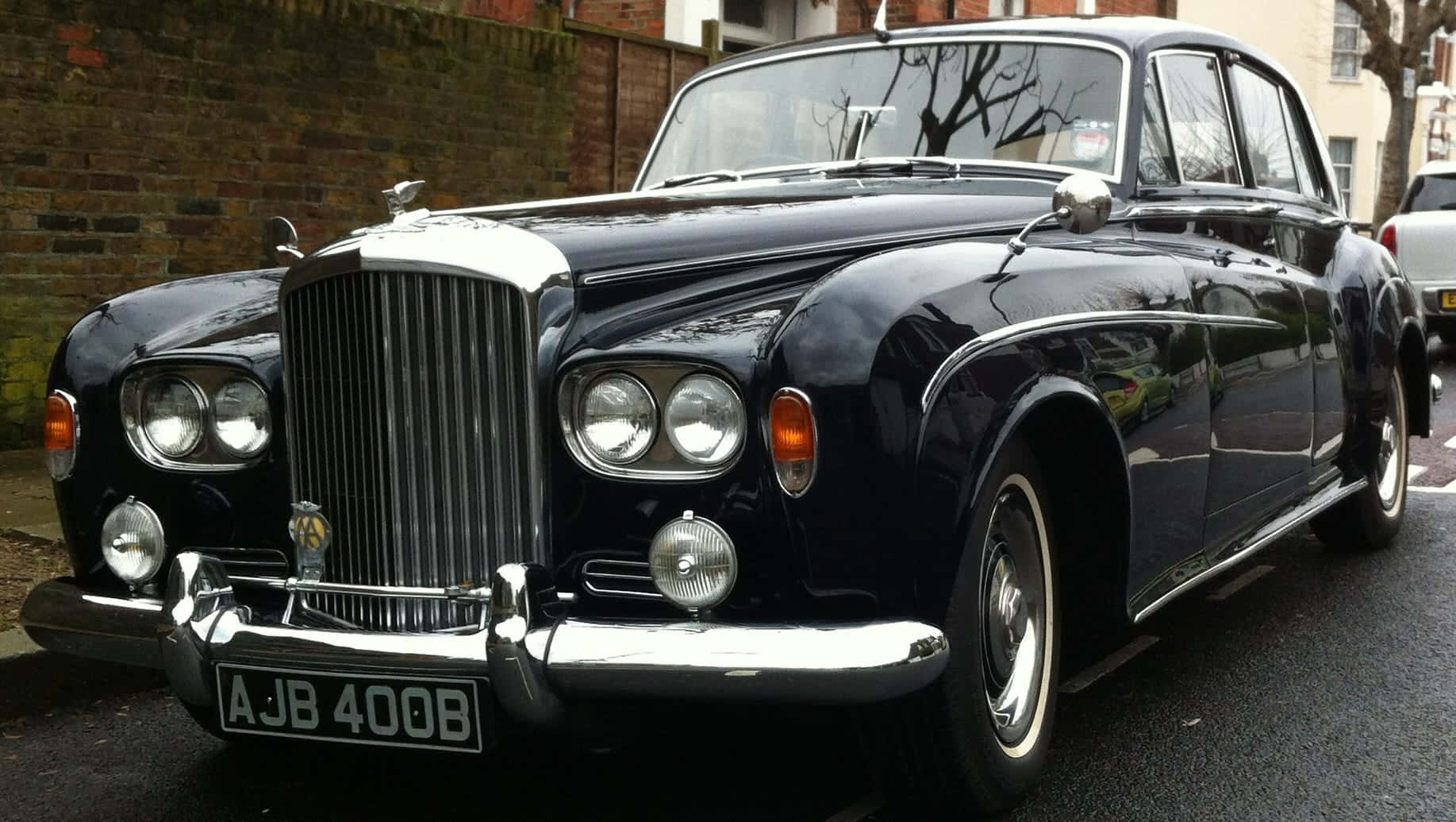 Bentley S3 桌布 壁紙 — 一輛亮黑色復古賓利，帶有鍍鉻裝飾，停在潮濕的城市街道上，旁邊是磚牆。