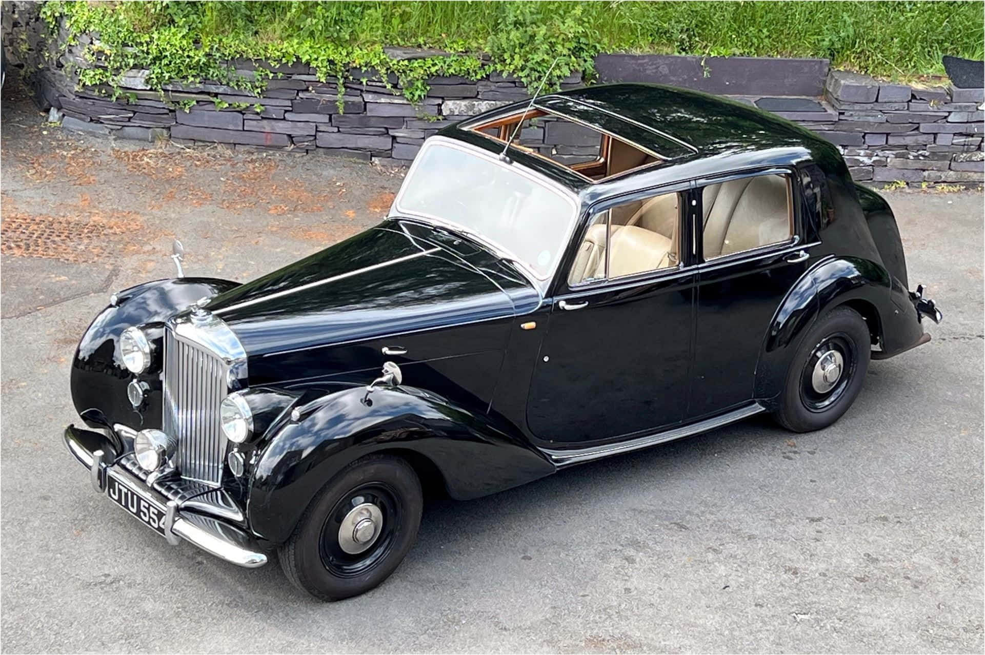 Bentley Mark Vi 桌布 壁紙 — 一架亮黑色 vintage Rolls-Royce 停泊在鋪砌路面上，旁邊係有綠色植物嘅石牆。