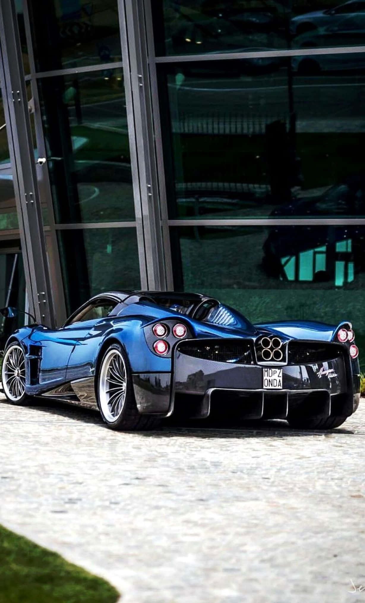 Pagani Huayra Roadster Bc 桌布 壁紙 — 一架閃亮藍色Pagani Huayra跑車停泊在現代玻璃建築旁的石鋪路面上。