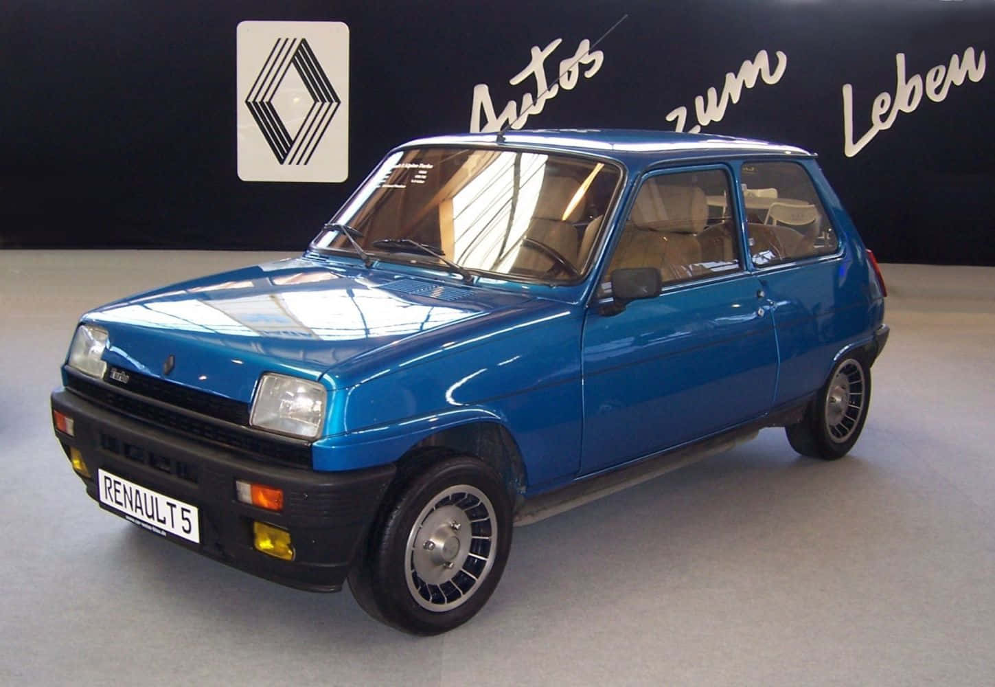 Renault 5 Turbo 桌布 壁紙 — 一輛閃亮的藍色雷諾5經典汽車在陳列室中展示，牆壁為黑色，帶有雷諾標誌和德語文字。