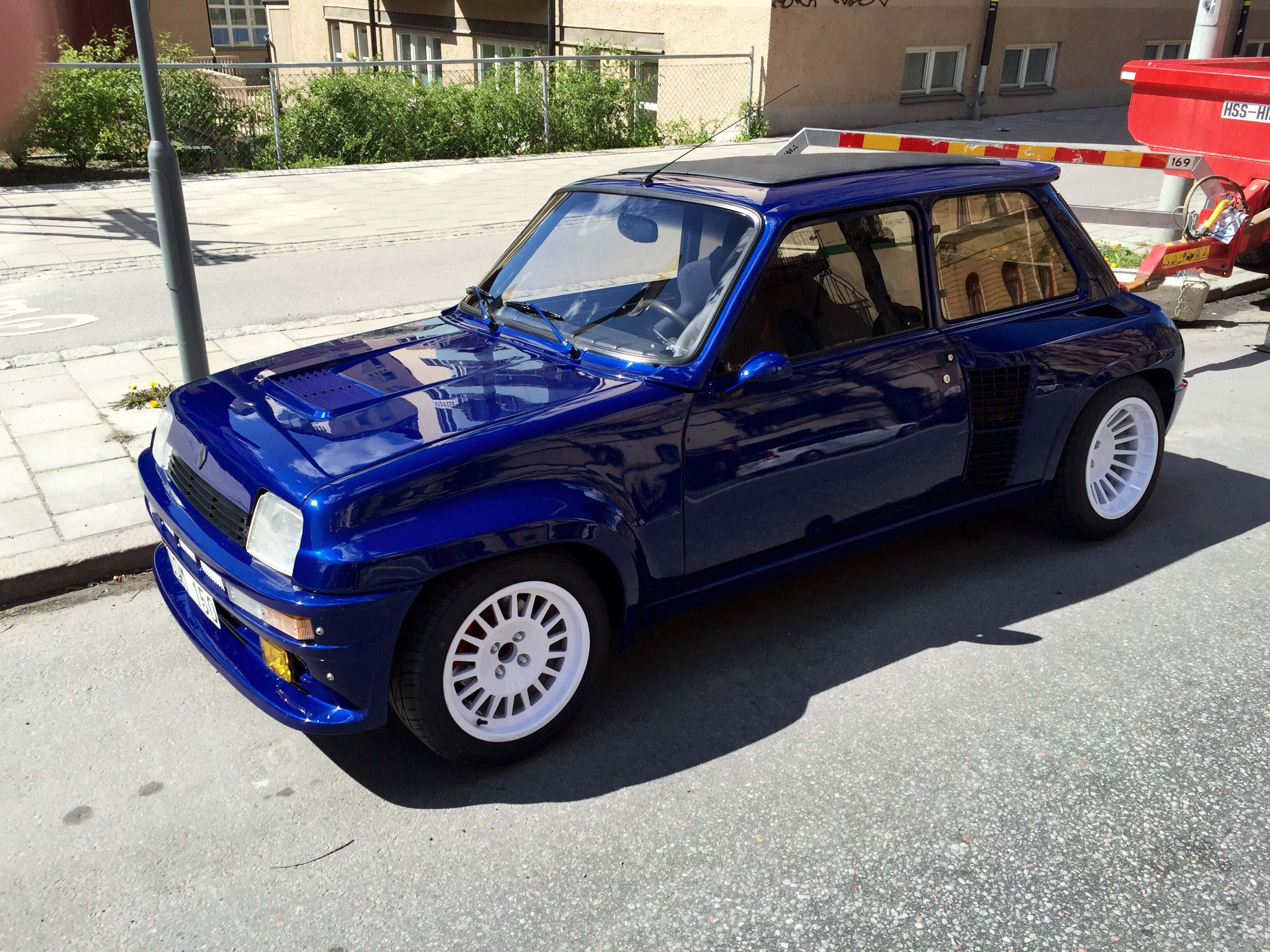 Renault 5 Turbo 桌布 壁紙 — 一輛光澤藍色雷諾5 Turbo跑車停泊在陽光明媚的城市街道上，配有白色車輪和附近的紅色卡車。