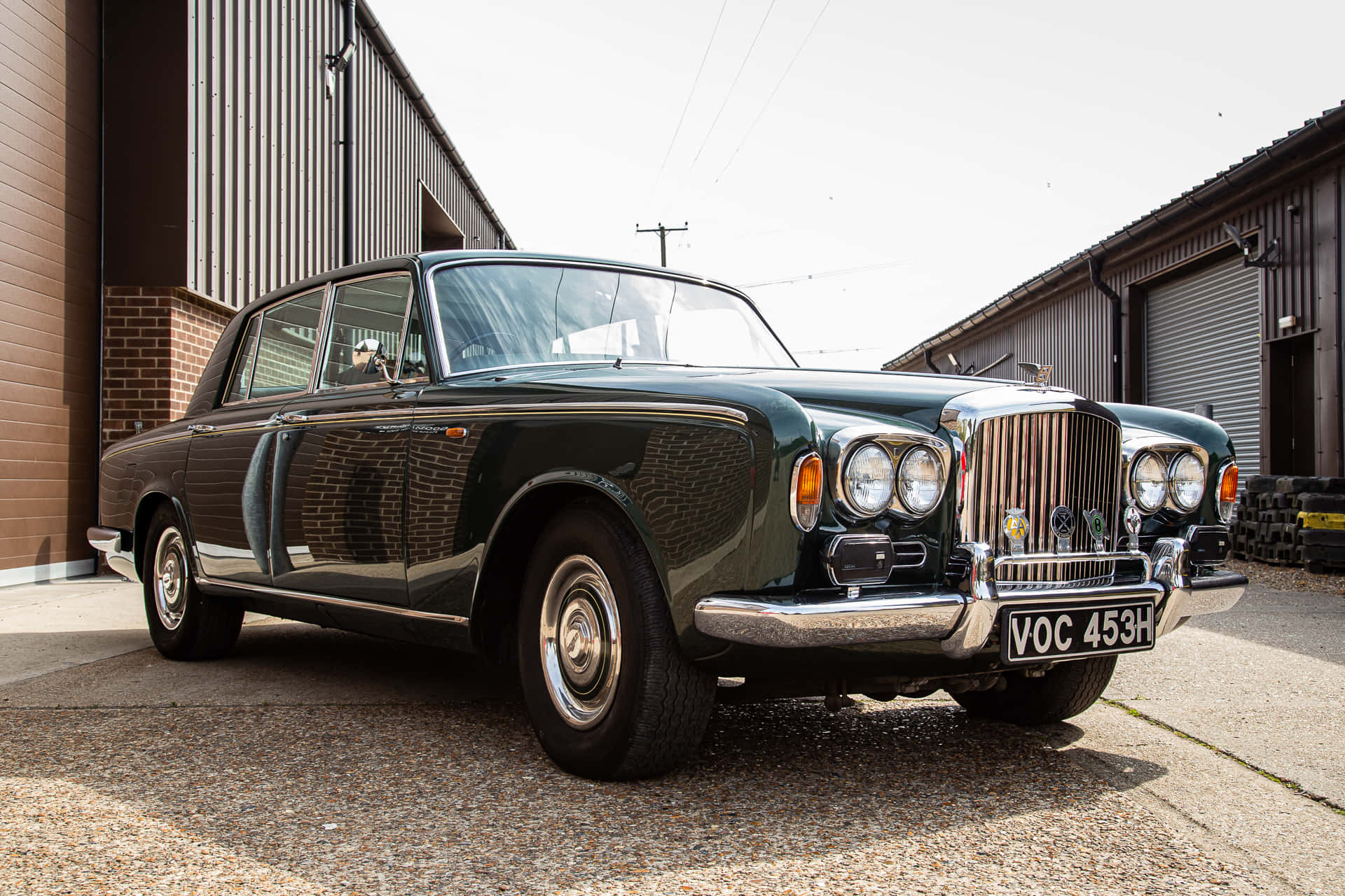 Bentley T1 桌布 壁紙 — 一輛光澤深綠色復古賓利轎車停泊在工業建築之間的礫石上，在明亮的日光下。