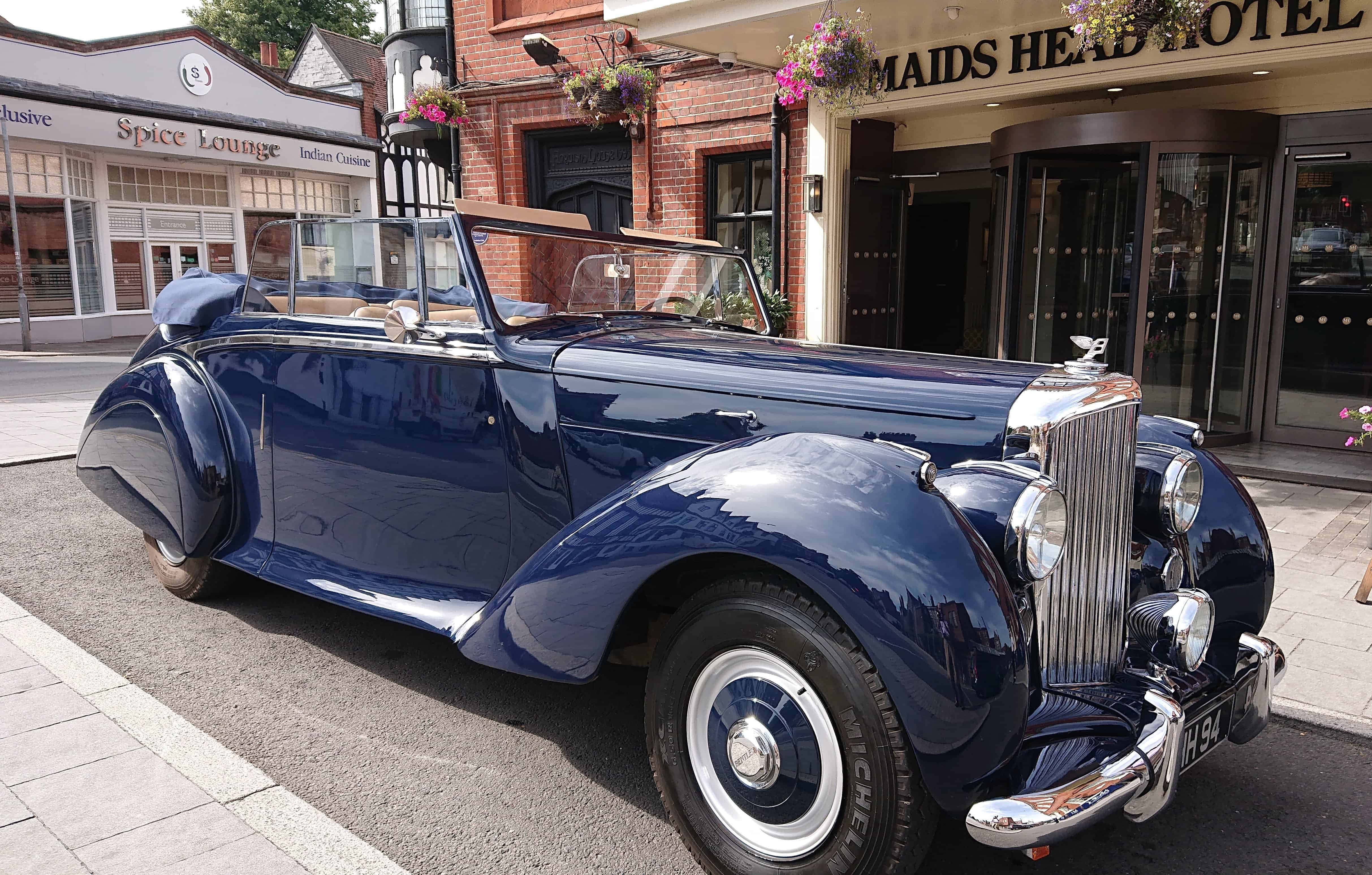 Bentley Mark Vi 桌布 壁紙 — 一架光澤深藍色復古開篷車停在鵝卵石街道上，前面係一間有掛花籃嘅磚砌酒店。