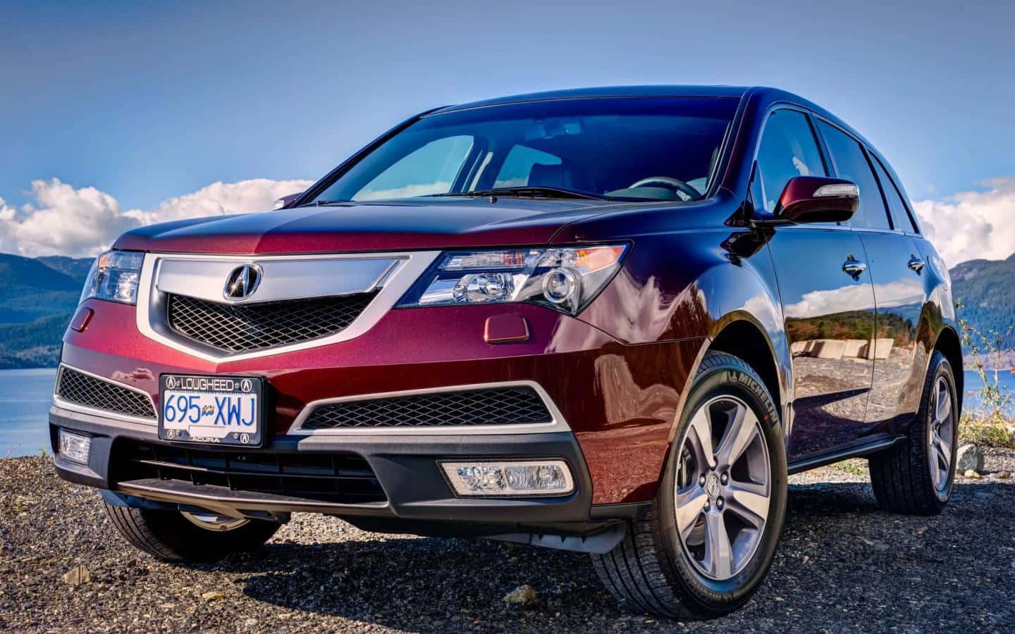 Acura Mdx 桌布 壁紙 — 一輛亮面酒紅色Acura SUV停泊在湖邊的碎石地上，背景有山巒和雲朵。