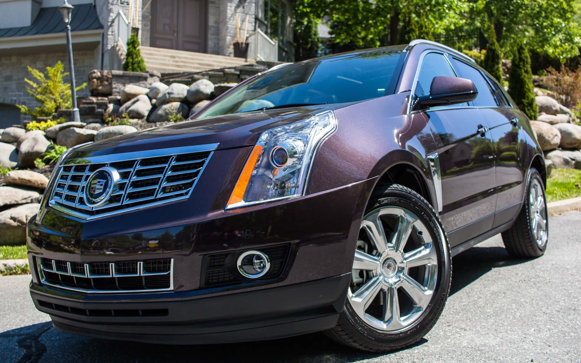 Cadillac Srx 桌布 壁紙 — 一輛光澤的栗色Cadillac SRX SUV停泊在陽光明媚的住宅街道上，周圍有石頭景觀和綠樹。