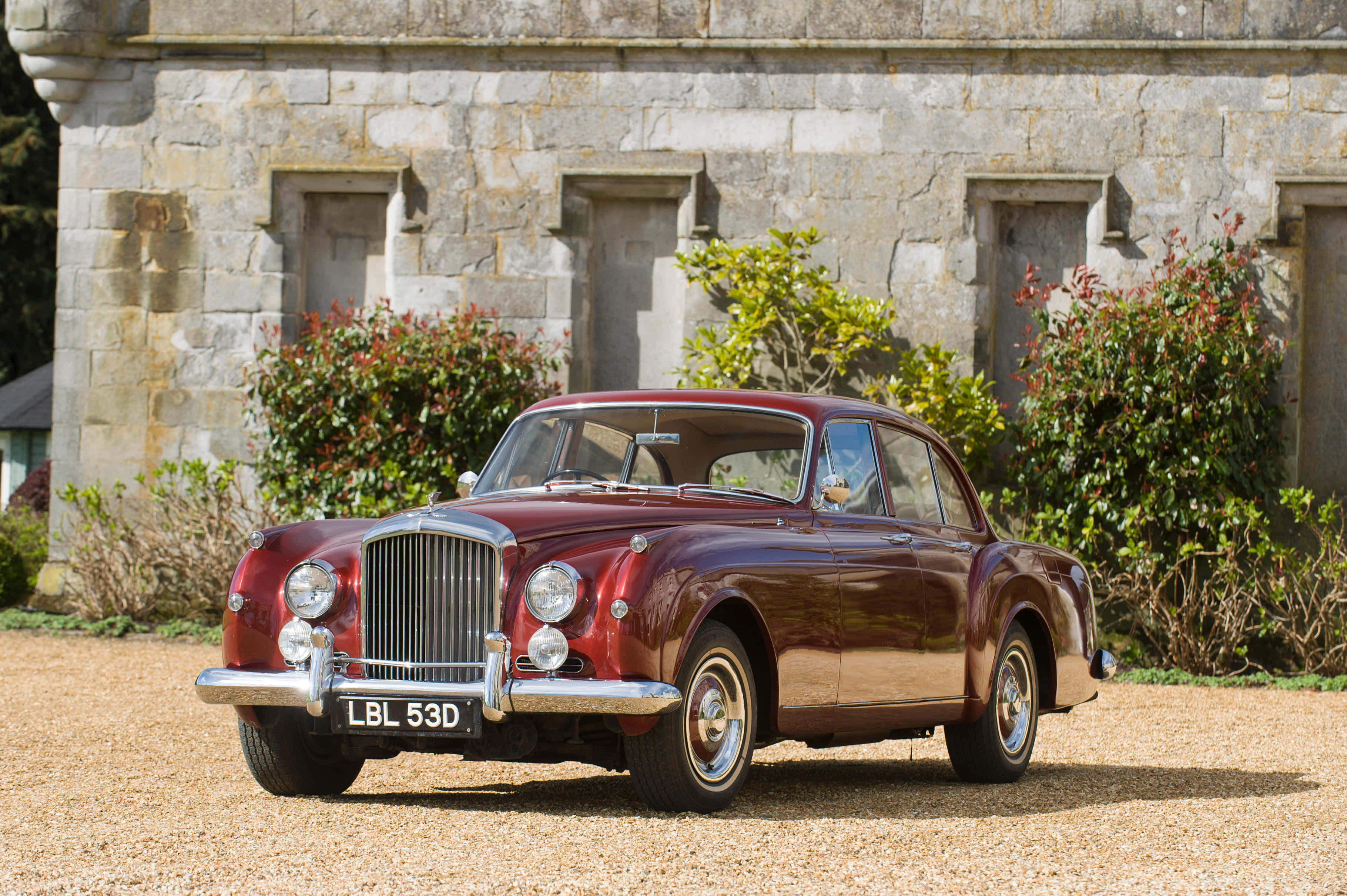 Bentley S2 桌布 壁紙 — 一架亮面栗色復古賓利房車停泊在碎石路上，前面係有綠色植物嘅石建築物。