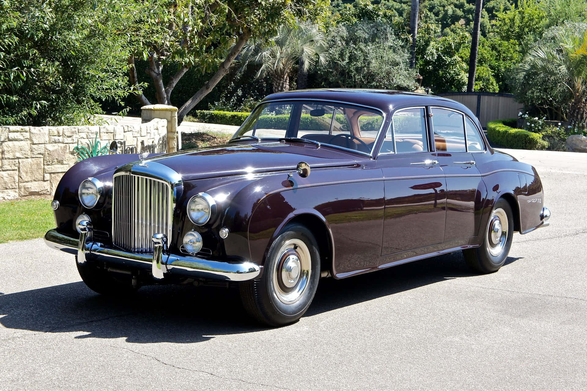 Bentley S2 桌布 壁紙 — 一輛光澤的栗色復古賓利轎車在陽光明媚的鋪砌車道上閃閃發光，周圍環繞著茂密的綠色植物。