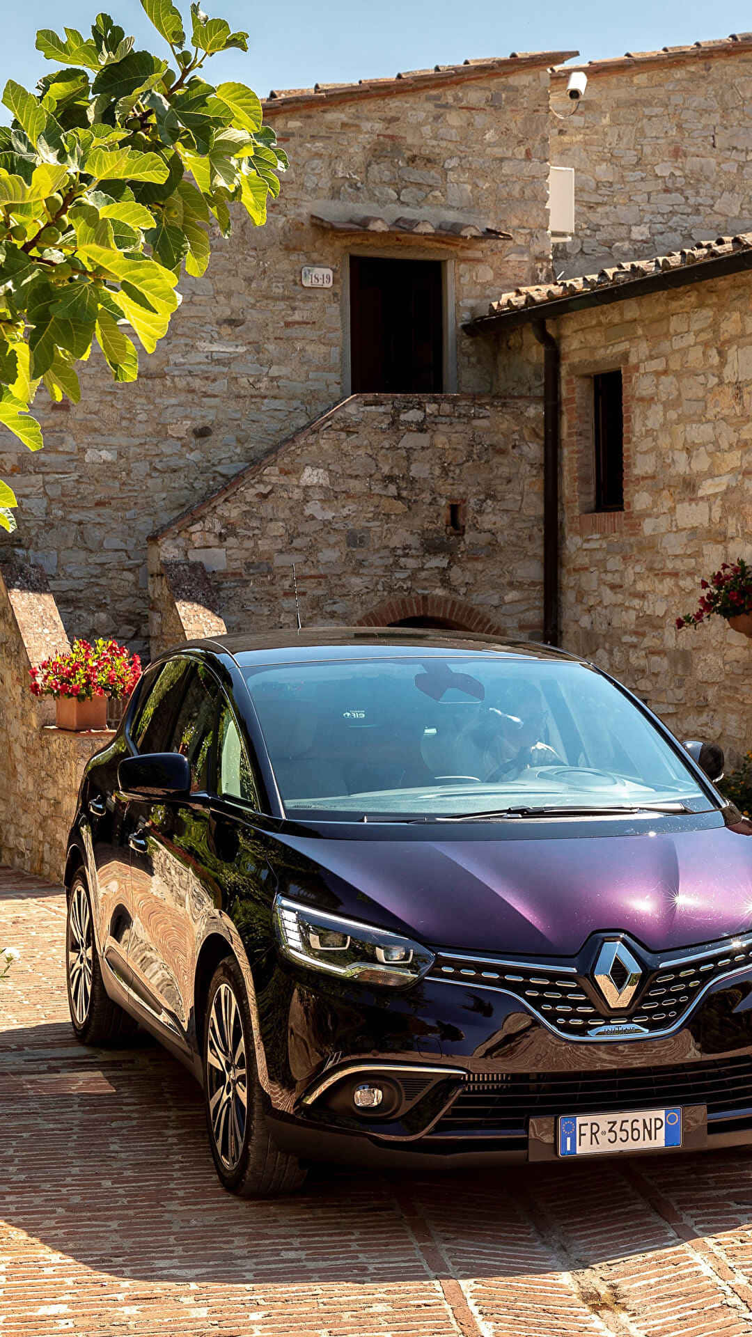 Renault Scenic 桌布 壁紙 — 一輛光澤的紫色雷諾 Koleos 停放在陽光明媚的鵝卵石庭院旁，旁邊是一座有綠色葉子的質樸石建築。