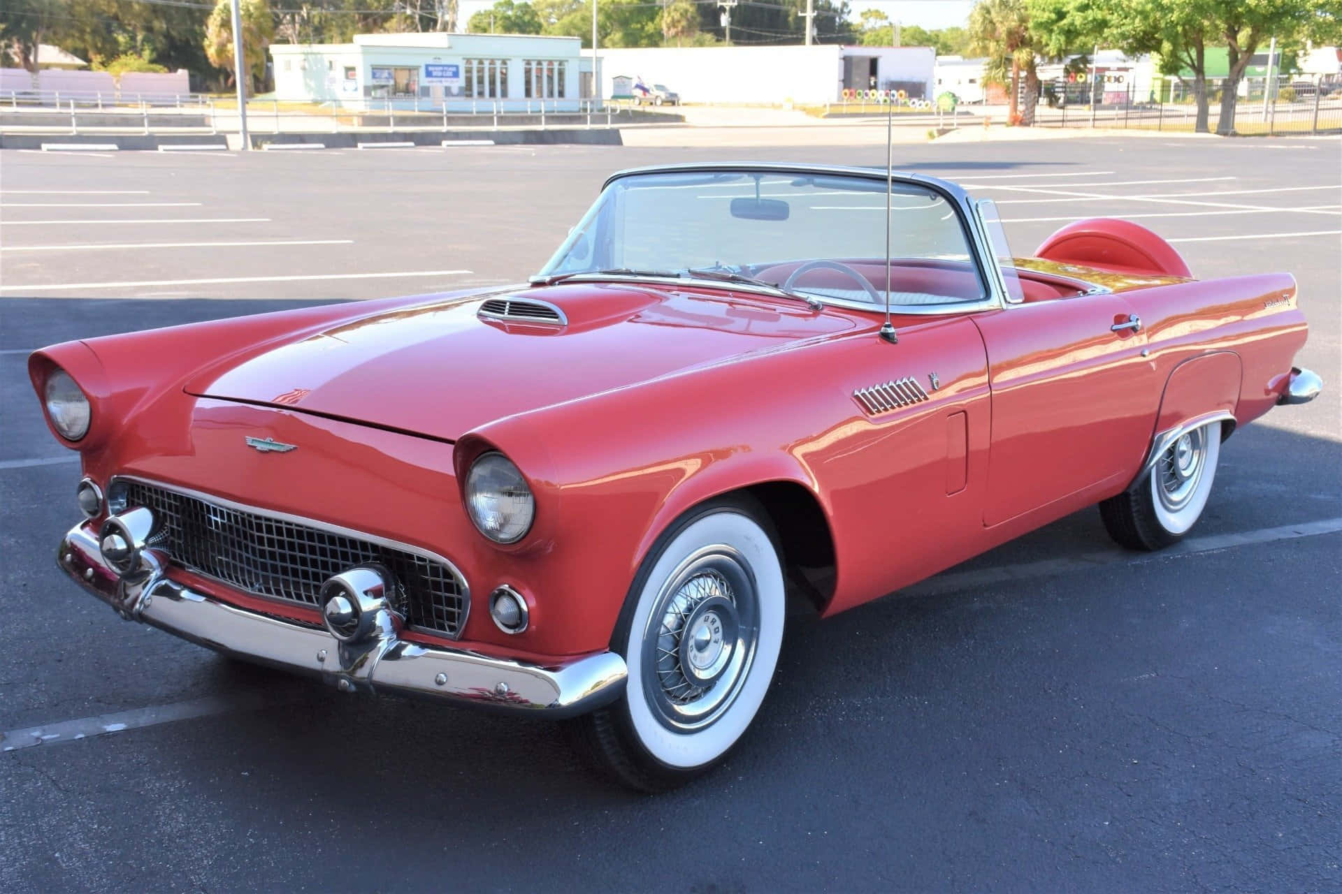 Ford Thunderbird 桌布 壁紙 — 一架閃亮的紅色1950年代福特雷鳥開篷車停泊在陽光明媚的瀝青停車場，帶有鍍鉻裝飾和白色輪胎壁。