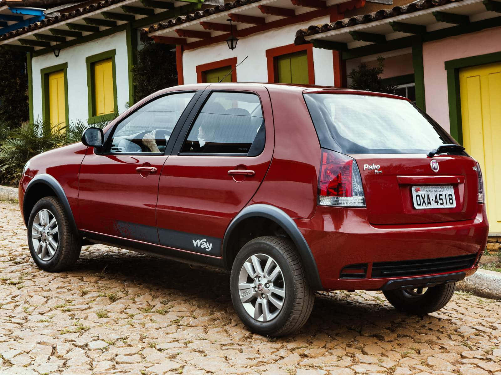 Fiat Palio 桌布 壁紙 — 一輛亮紅色的Fiat Punto Way停泊在陽光普照的鵝卵石車道上，前面是一座色彩繽紛的殖民風格房屋。