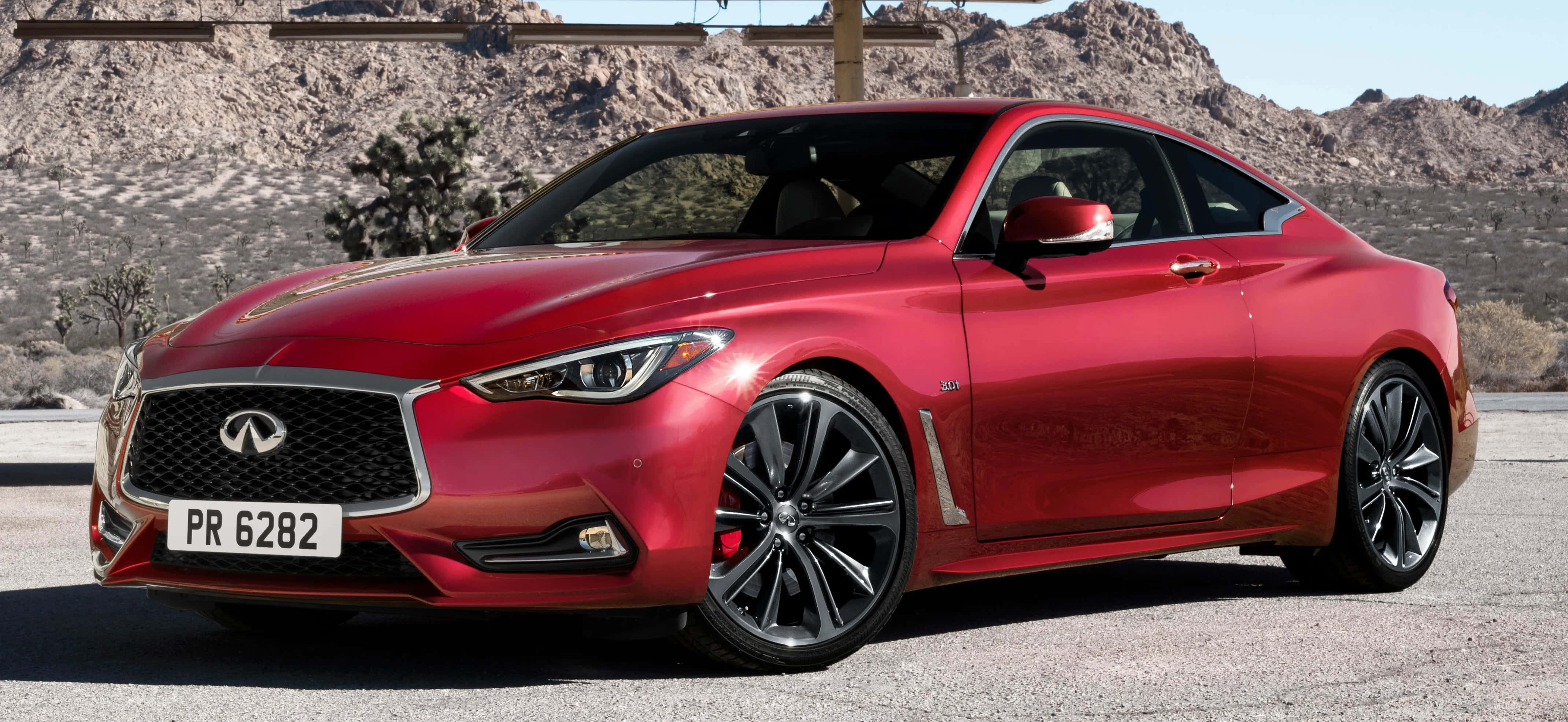 Infiniti Q60 桌布 壁紙 — 一輛亮紅色的Infiniti Q60運動轎跑車停在瀝青路上，背景是岩石沙漠山脈和稀疏樹木，在明亮的日光下。