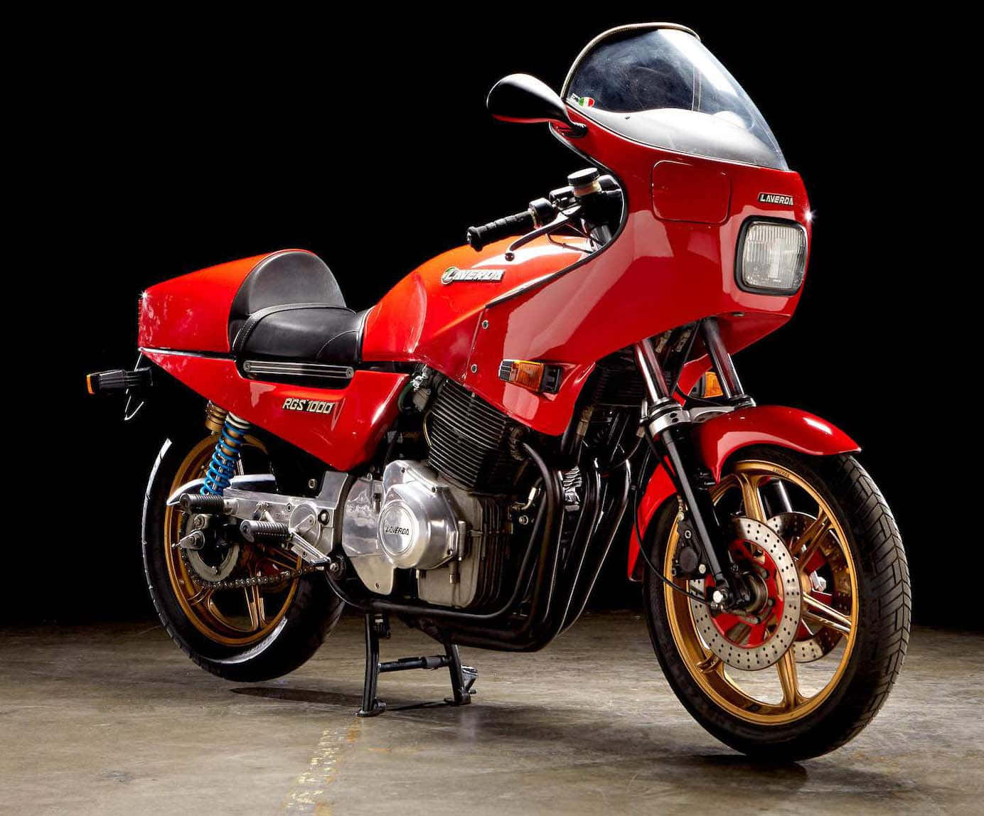  壁紙 — 一架光澤紅色 Kawasaki ZGS 1000 電單車，配金色輪圈和藍色懸掛，以側腳架停放在純黑背景前。