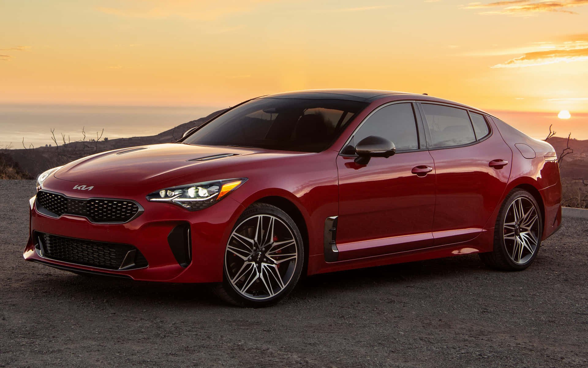 Kia Stinger 桌布 壁紙 — 一架亮紅色嘅Kia Stinger運動房車泊喺日落時分嘅碎石觀景點，背景係橙色天空同遠處山丘。