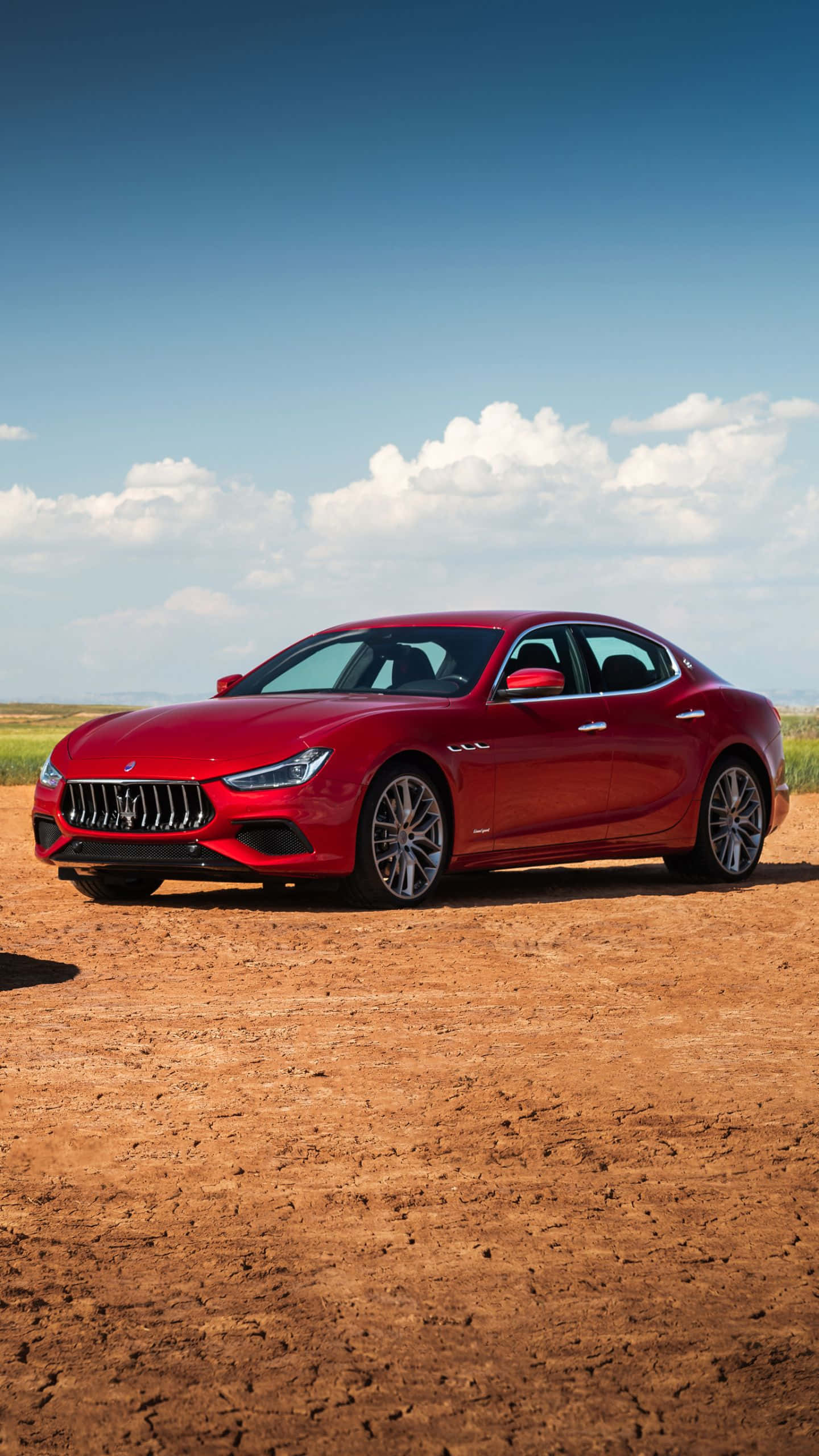Maserati Ghibli 桌布 壁紙 — 一輛亮紅色瑪莎拉蒂跑車停在龜裂的土地上，背景是晴朗的藍天和蓬鬆的白雲。
