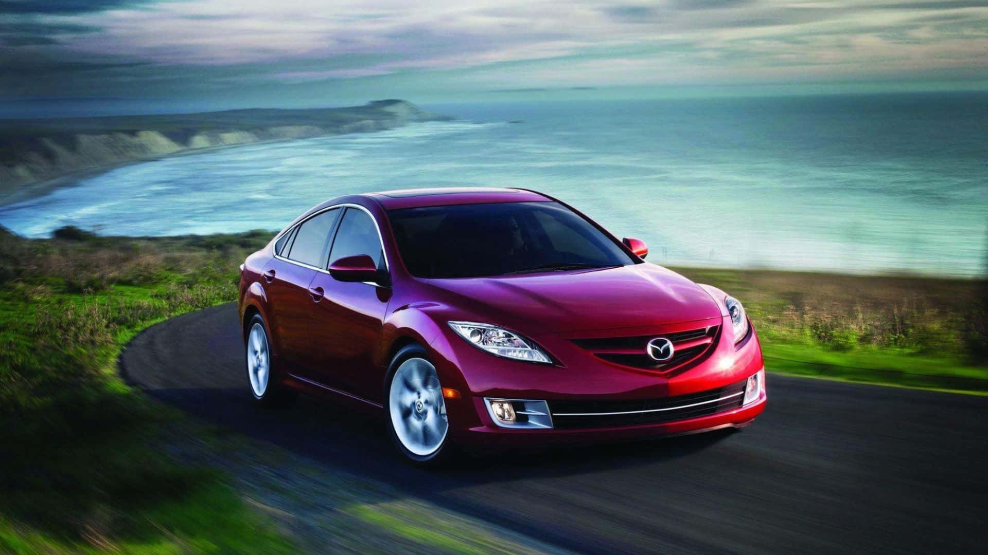 Mazda 6 桌布 壁紙 — 一輛亮紅色Mazda房車沿著海岸公路飛馳，背景是模糊的綠色山丘和碧綠色的海洋。