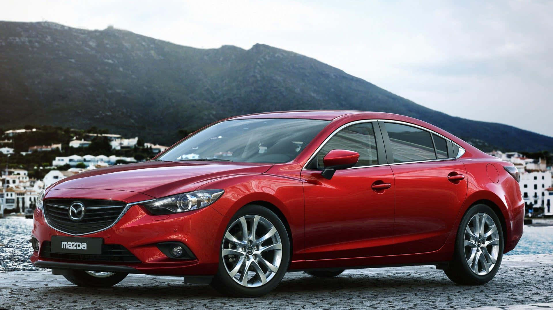 Mazda 6 桌布 壁紙 — 一架亮紅色Mazda房車停泊在鵝卵石海濱，背景有山脈和白色建築物。