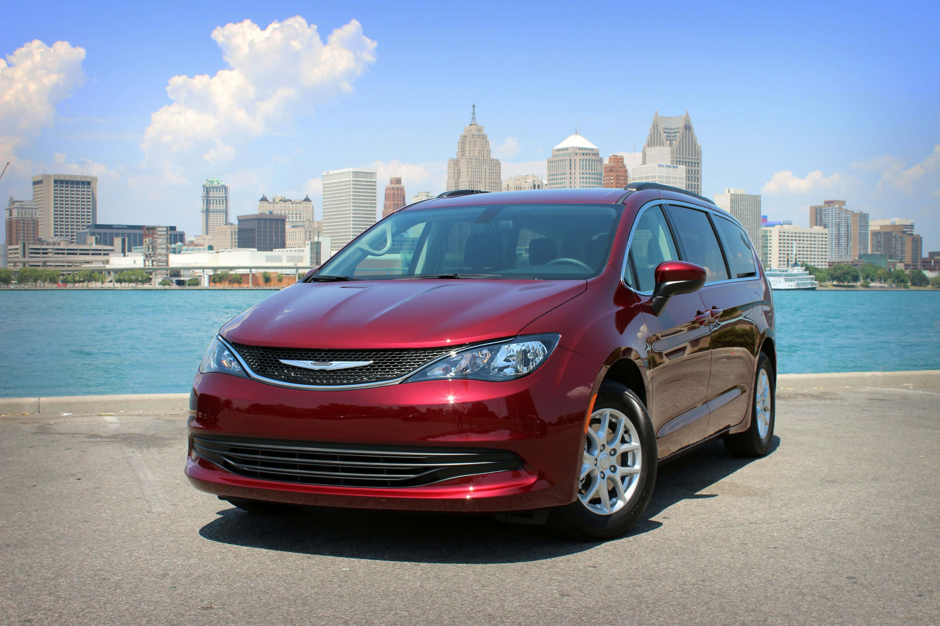 Chrysler Voyager 桌布 壁紙 — 一輛光亮的紅色小型貨車停在水邊，城市天際線在明亮藍天和蓬鬆白雲下。