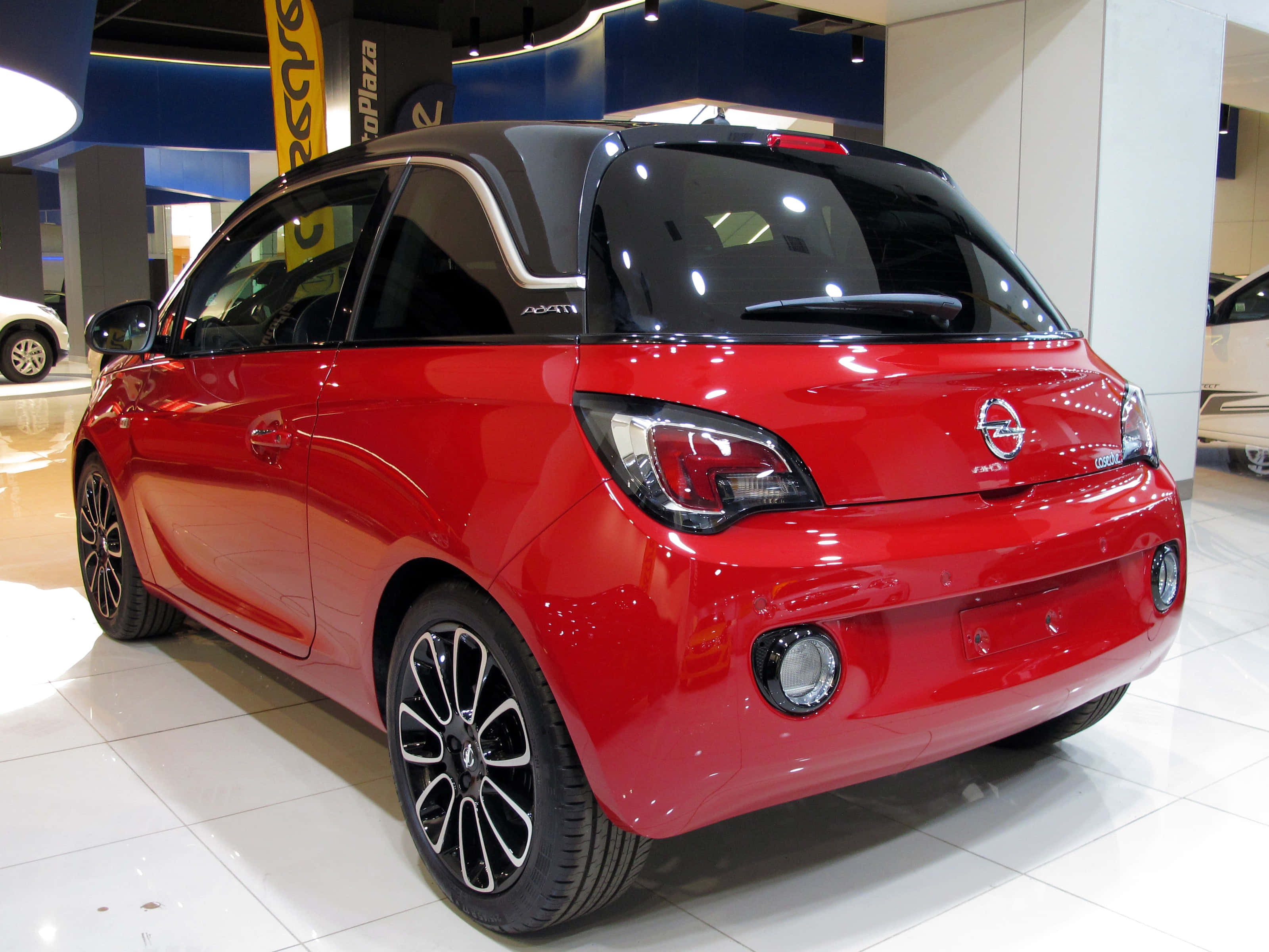 Opel Adam 桌布 壁紙 — 一輛光亮的紅色Opel Adam緊湊型汽車在明亮的室內汽車展廳中從後方展示，地板鋪有瓷磚並帶有品牌標誌。