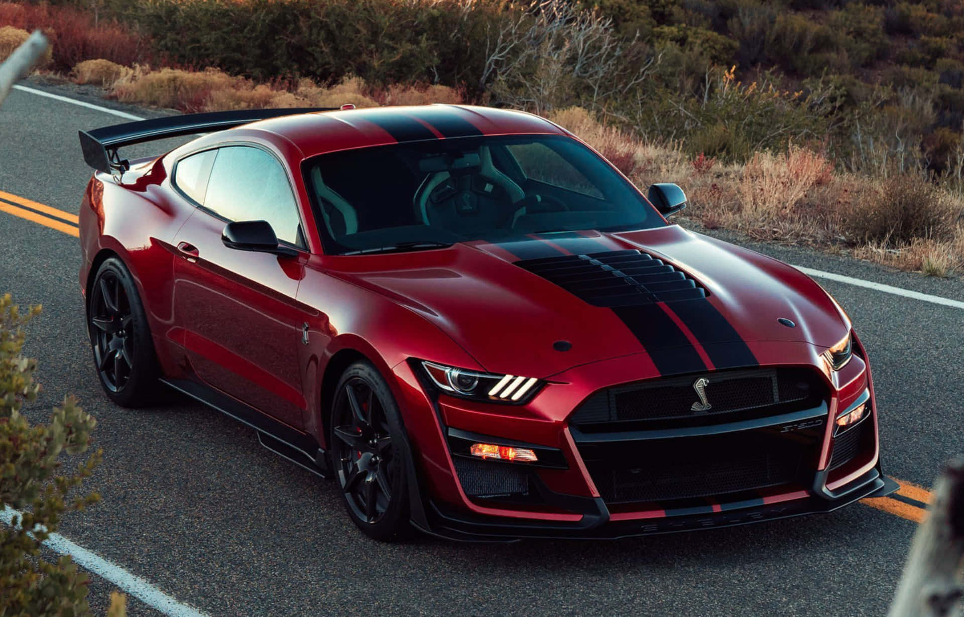  壁紙 — 一輛帶有黑色賽車條紋的亮紅色Shelby GT500跑車在日落時分沿著蜿蜒的沙漠公路行駛。