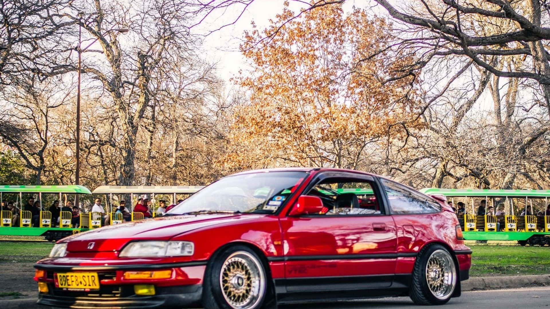 Honda Civic Del Sol 桌布 壁紙 — 一輛光亮的紅色跑車停泊在公園內，背景有光禿的樹木和綠色電車。