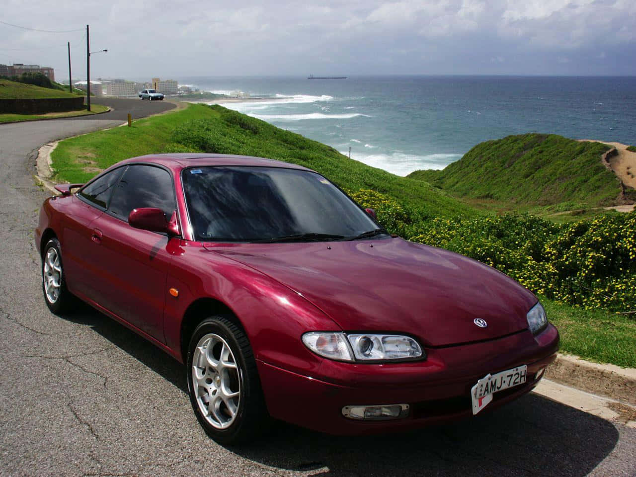 Mazda Mx-6 桌布 壁紙 — 一架亮紅色跑車停泊在沿海公路上，背景有綠色山丘和洶湧的海浪。