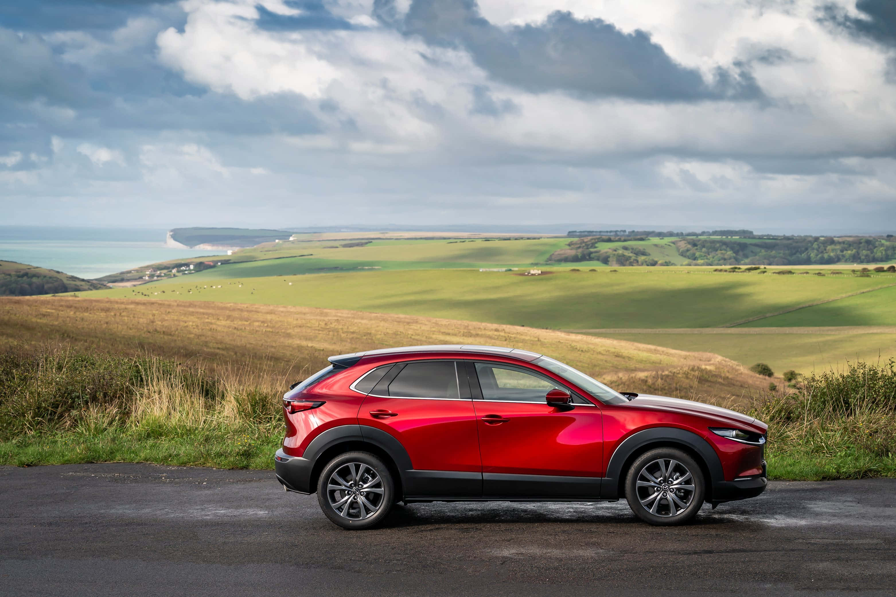 Mazda Cx-30 桌布 壁紙 — 一架亮紅色SUV停泊喺沿海公路，俯瞰住起伏嘅綠色山丘同遠處嘅白色懸崖，天空多雲。