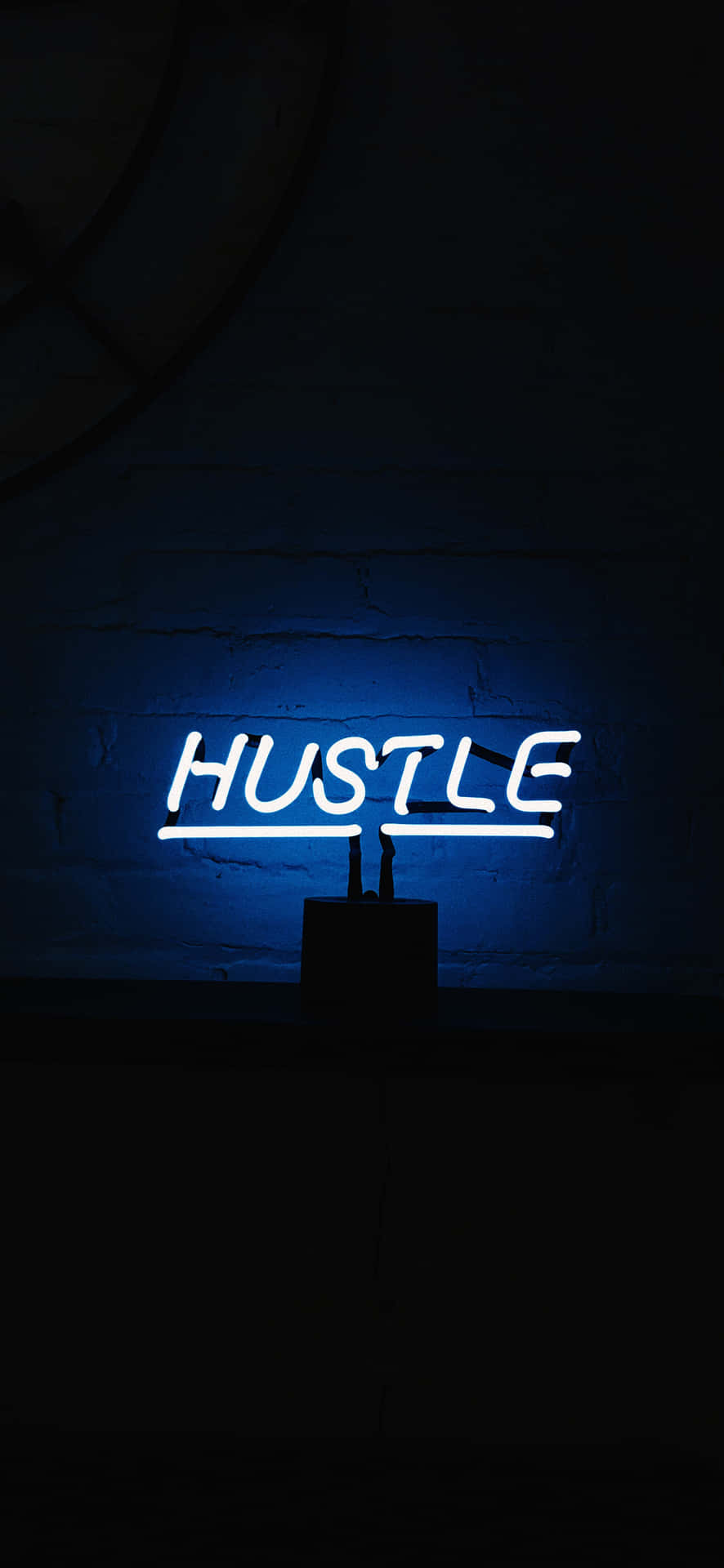  壁紙 — 一個發光的藍色霓虹燈牌，拼寫著「HUSTLE」，照亮了陰暗、簡約環境中的深色磚牆。