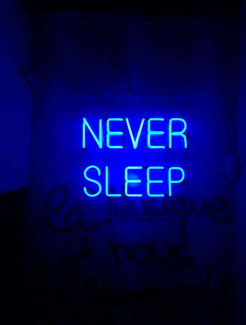  壁紙 — 一個發光的藍色霓虹燈牌，上面寫著「NEVER SLEEP」，在下方帶有模糊塗鴉的暗牆上投射出詭異的光芒。
