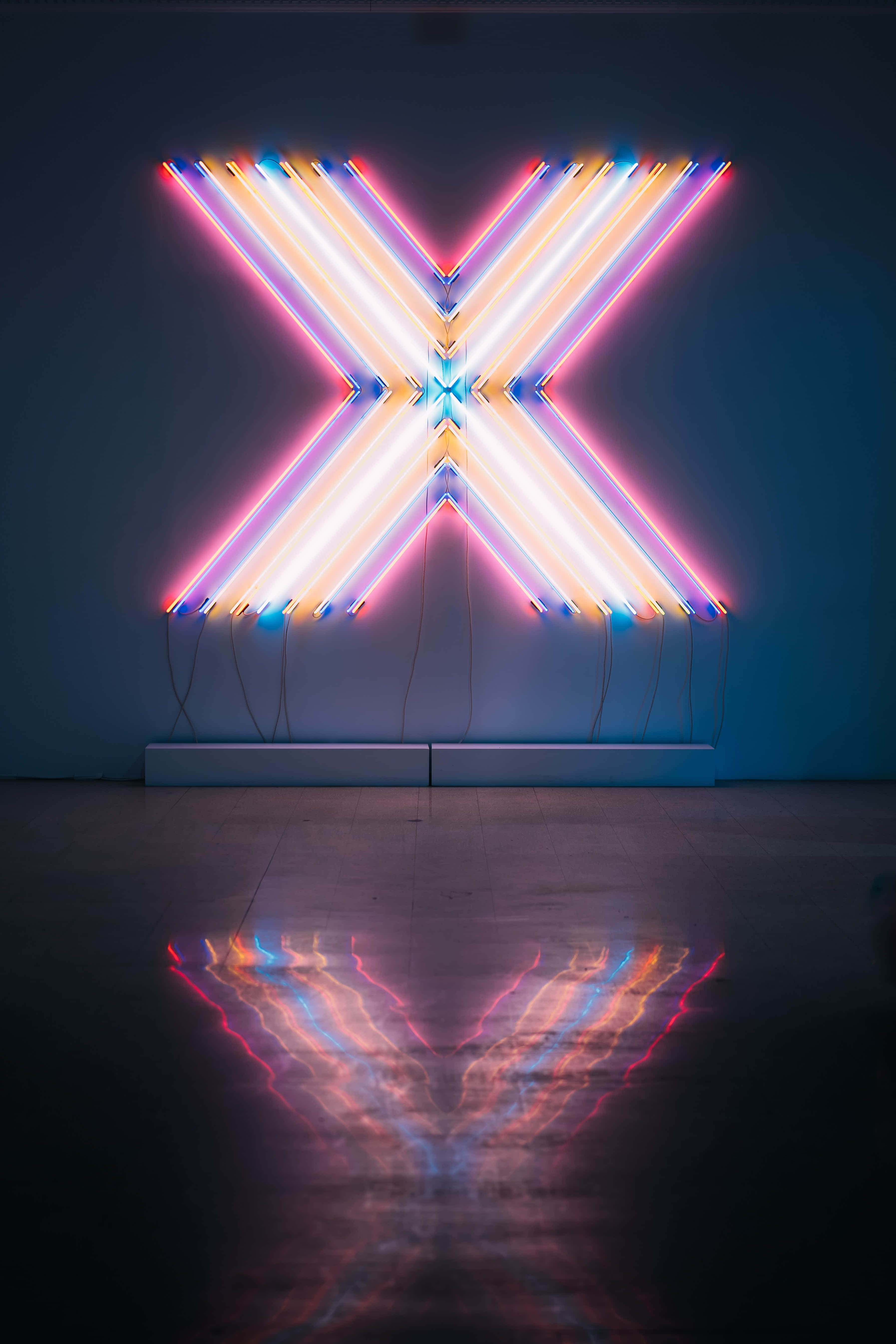 Neon Light 桌布 壁紙 — 一個發光的粉紅、藍色和白色霓虹燈X雕塑，在極簡主義畫廊空間的黑暗地板上投射出鮮豔的倒影。