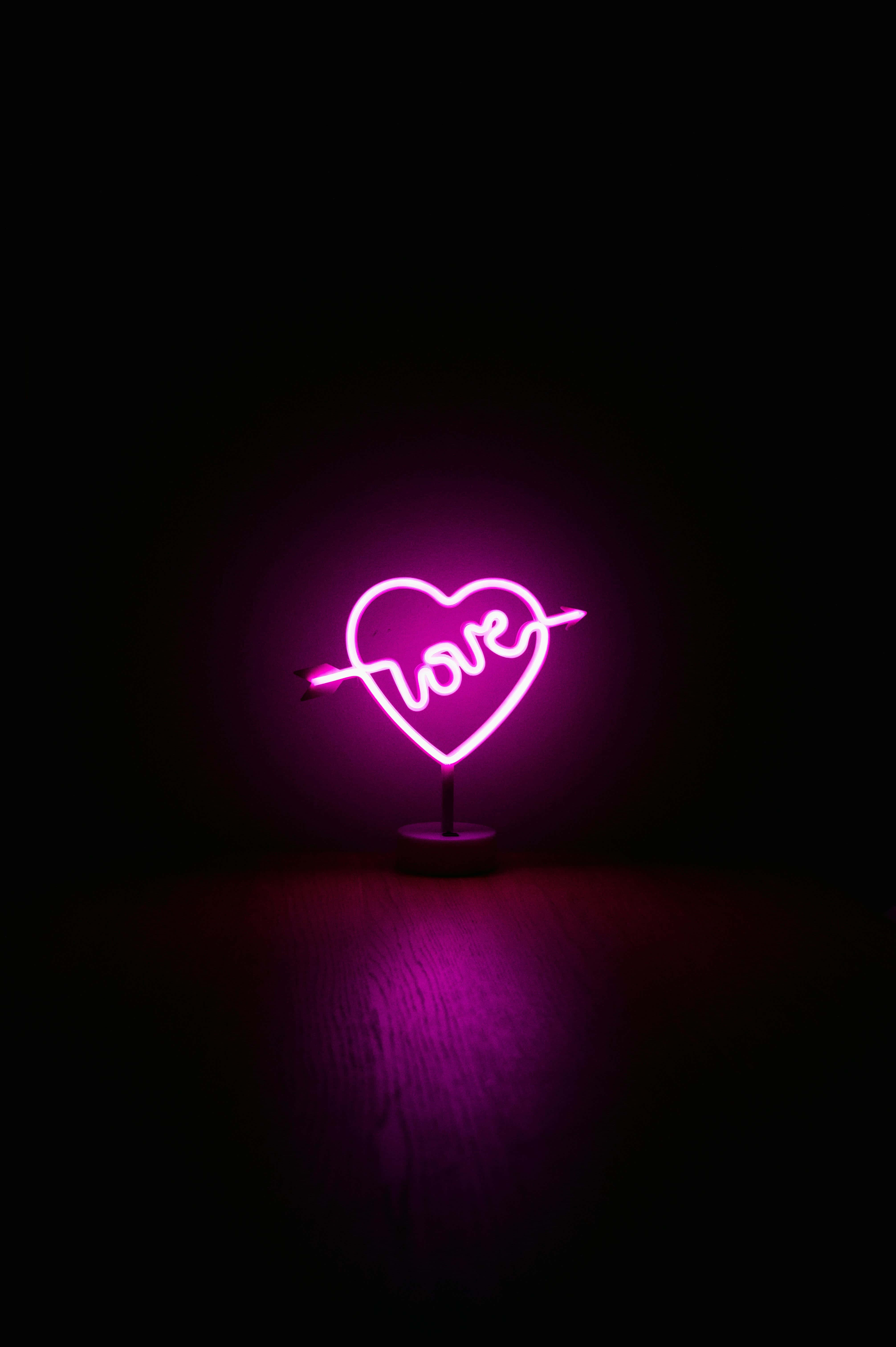 Neon Lights iPhone 桌布 壁紙 — 一個發光的粉紅色霓虹燈牌，形狀為被箭穿過的心形，內有「love」字樣，在深色木質表面上投射出柔和的光芒。