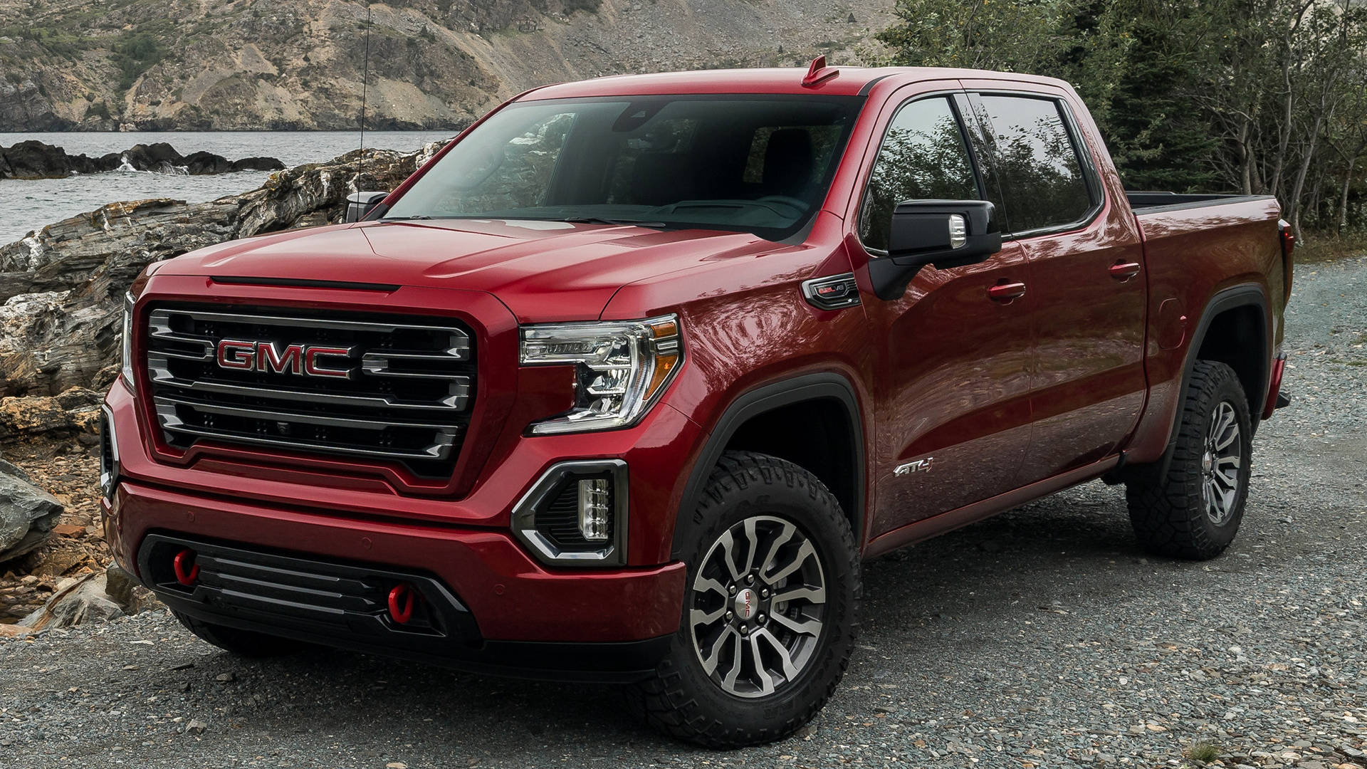 GMC Sierra 旁湖畔 壁紙 — 一輛亮紅色的GMC Sierra AT4皮卡車停在碎石上，靠近岩石海岸線，背景有水和樹木。