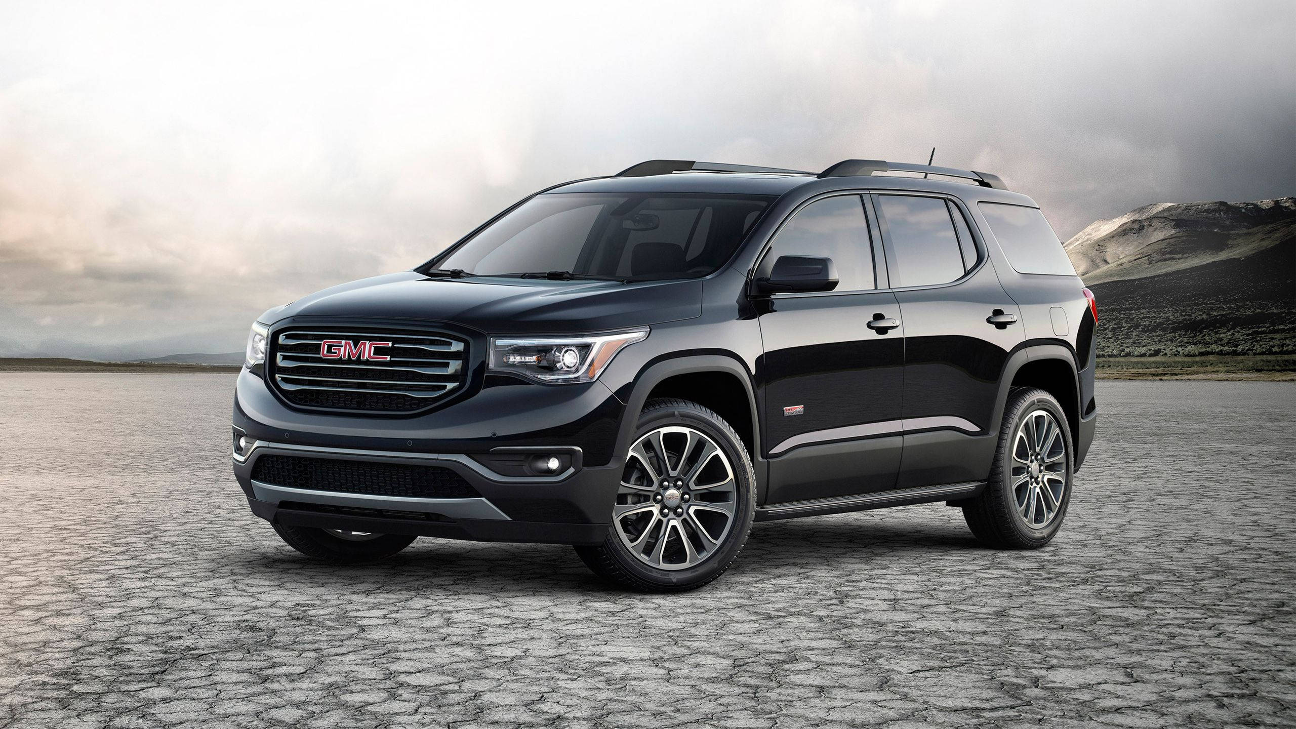 GMC 在崎嶇路徑上 壁紙 — 一架時尚的黑色GMC Acadia SUV停泊在龜裂的沙漠地面上，天空多雲，遠山在望。