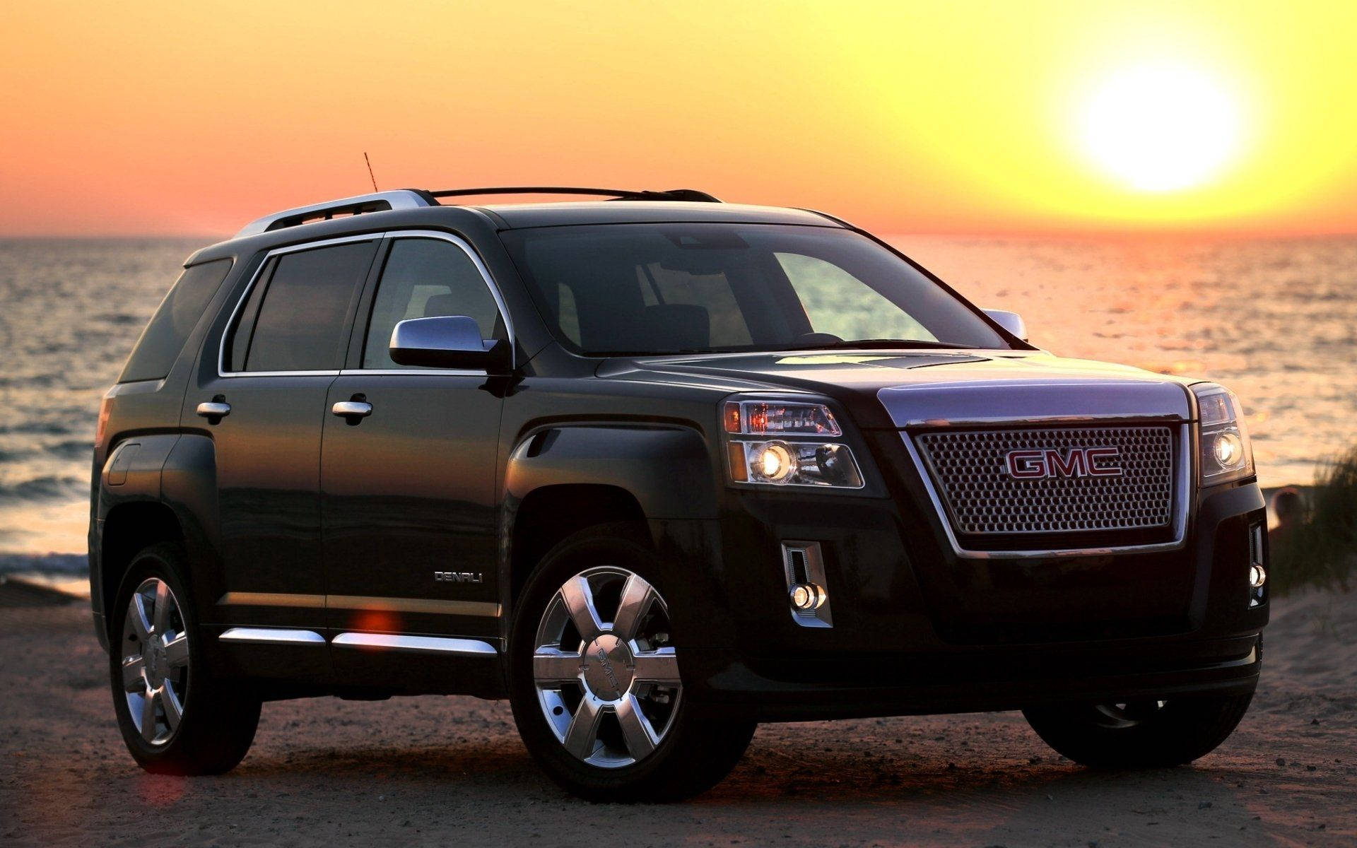 GMC汽車在晴朗日光下 壁紙 — 一輛時尚的黑色GMC Acadia SUV停在日落時分的沙灘上，金色光線反射在它的鉻合金輪圈和格柵上。