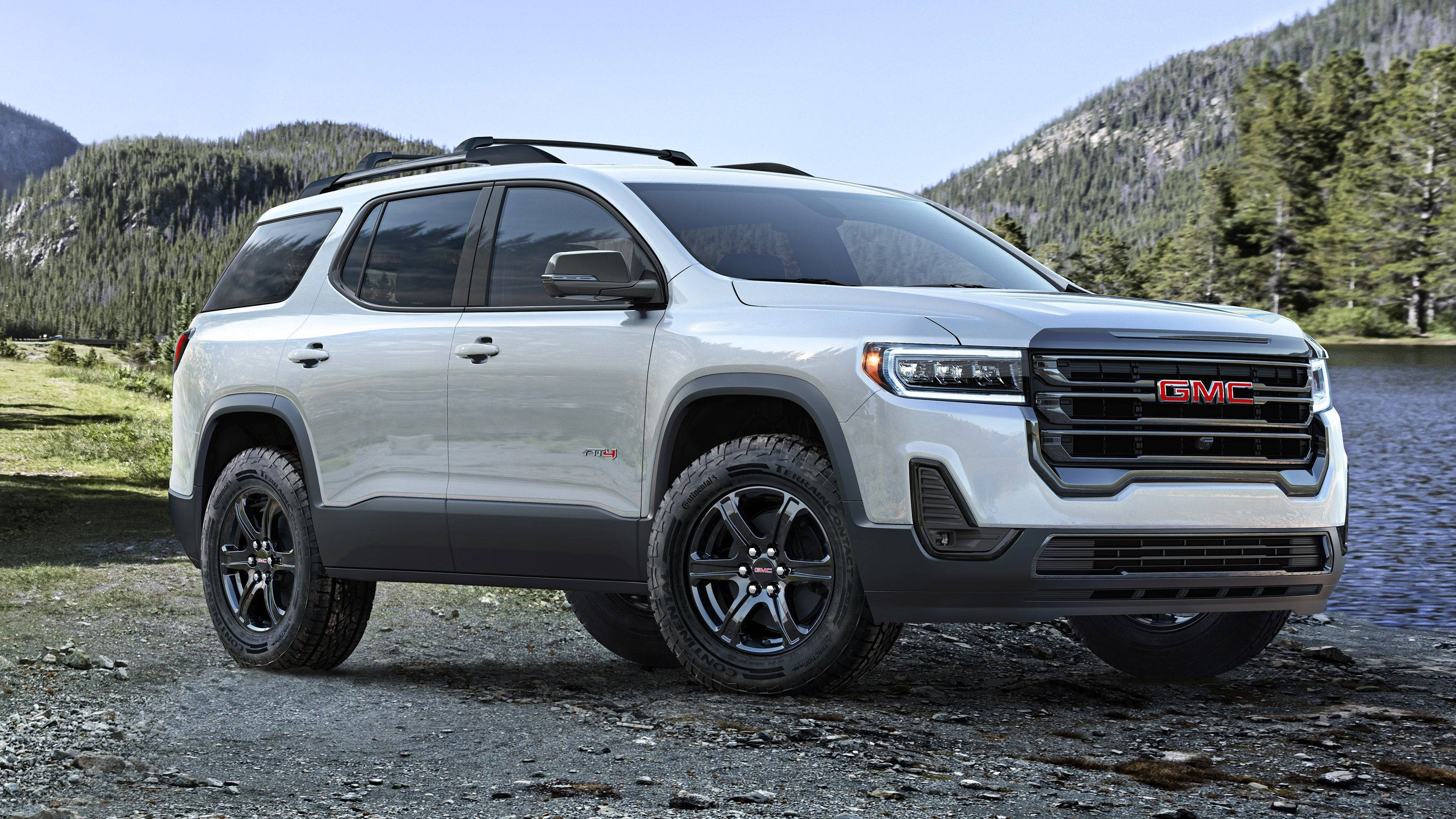 GMC白色Acadia車款 壁紙 — 一輛時尚的銀色GMC SUV，帶有黑色飾邊，停在平靜湖泊旁的礫石岸邊，背景是森林覆蓋的山脈。
