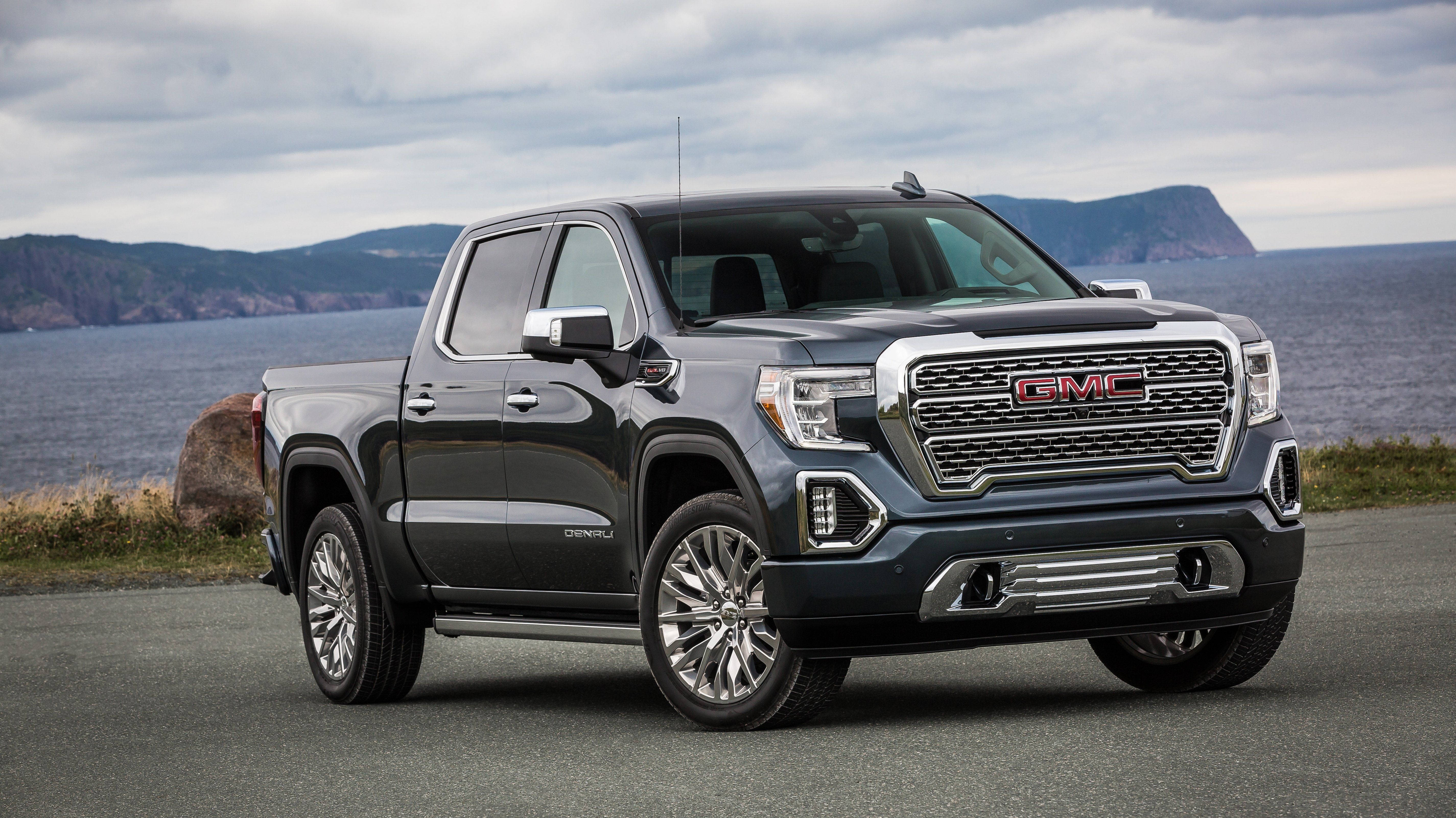 GMC車輛近海 壁紙 — 一輛時尚的深灰色GMC Sierra Denali皮卡車停泊在海岸公路上，背景是海洋和遠處的懸崖，天空多雲。