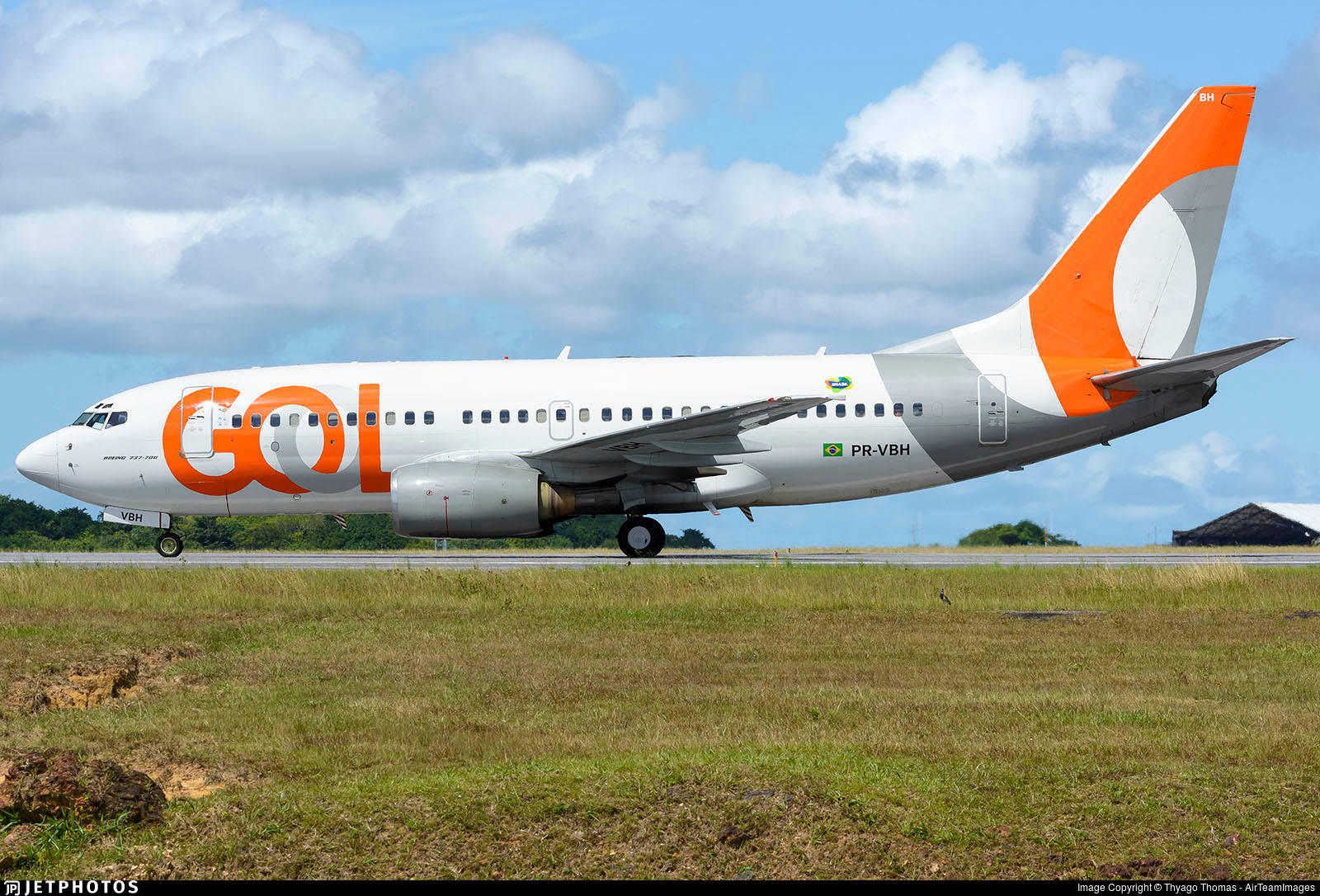 Gol 航空公司飛機停靠綠草地旁 壁紙 — 一架白色和橙色的戈爾航空波音737在部分多雲的藍天下，在跑道滑行，前景有綠色草地。