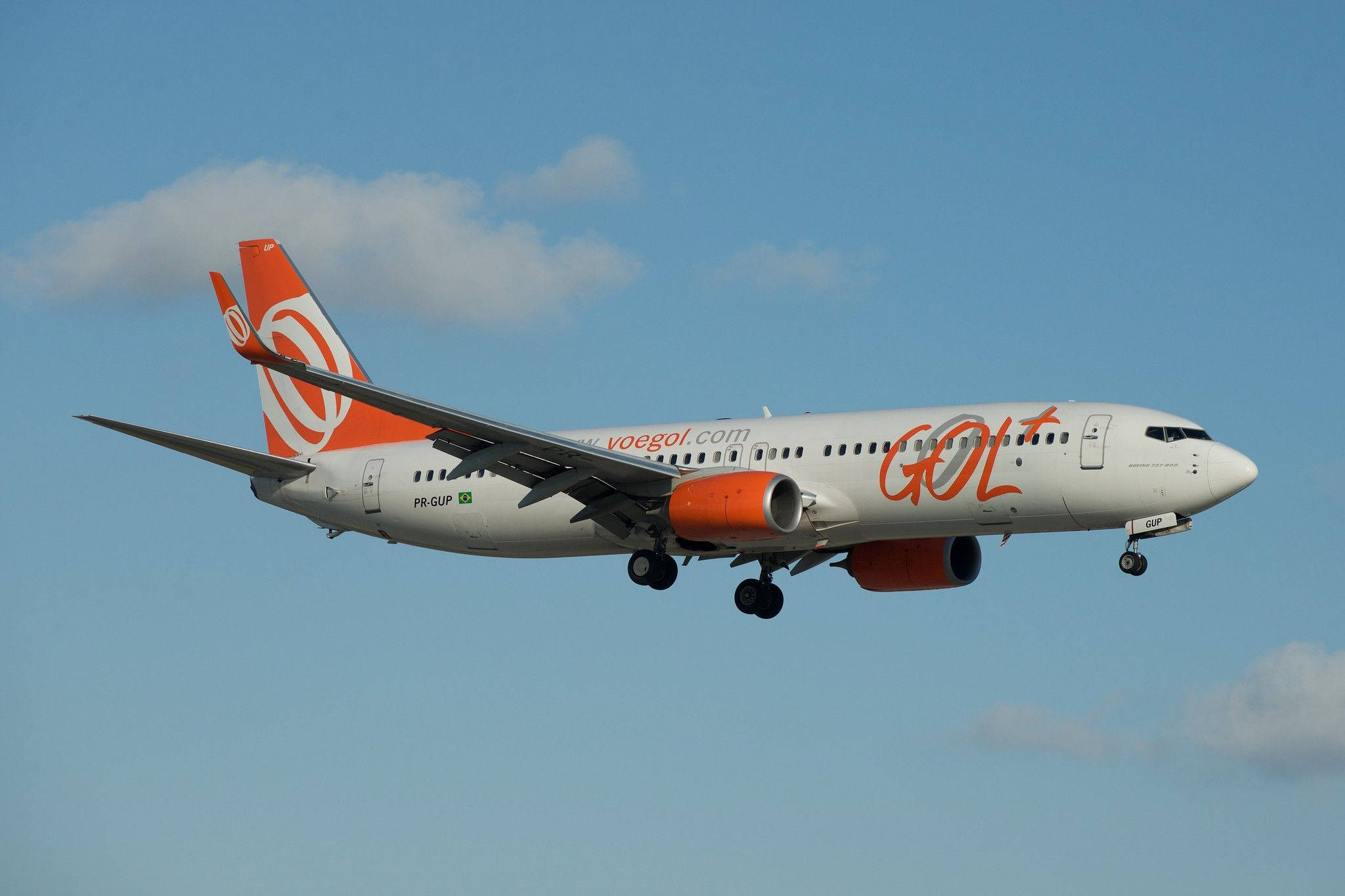 Gol 航空公司飛機翱翔半空 壁紙 — 一架白色和橙色嘅Gol Airlines波音737，起落架放下，喺晴朗藍天同零星雲朵中翱翔。
