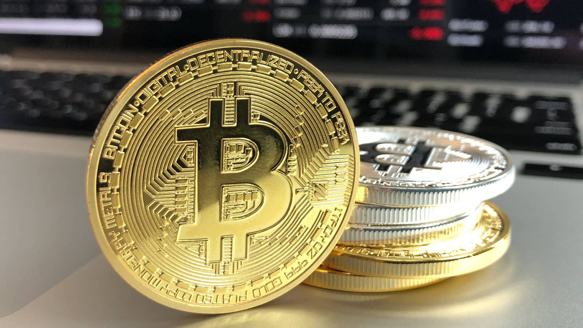 一堆實體比特幣硬幣放在筆記型電腦鍵盤上，後面有金融圖表。