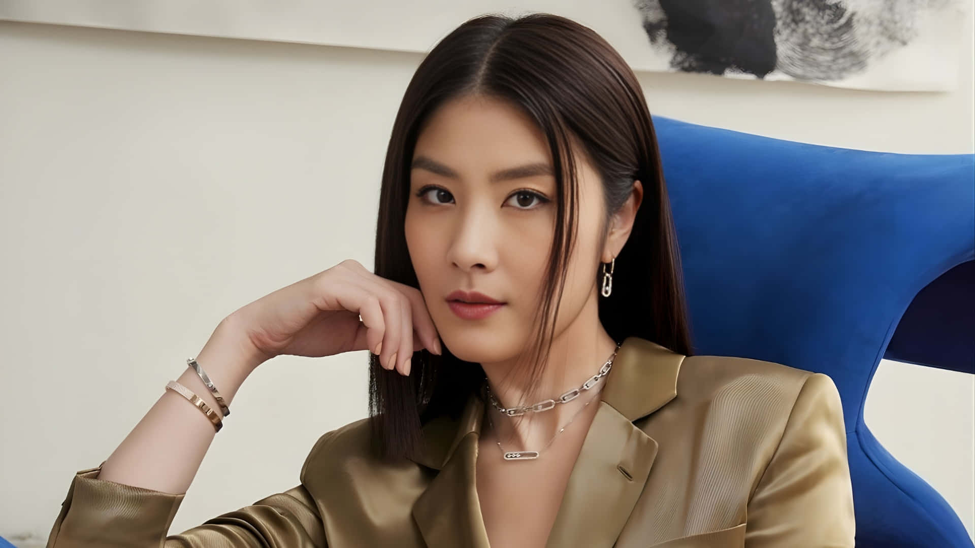  壁紙 — 一位身穿金色西裝外套的優雅女士，在鮮藍色椅子前托腮，背後是抽象藝術。