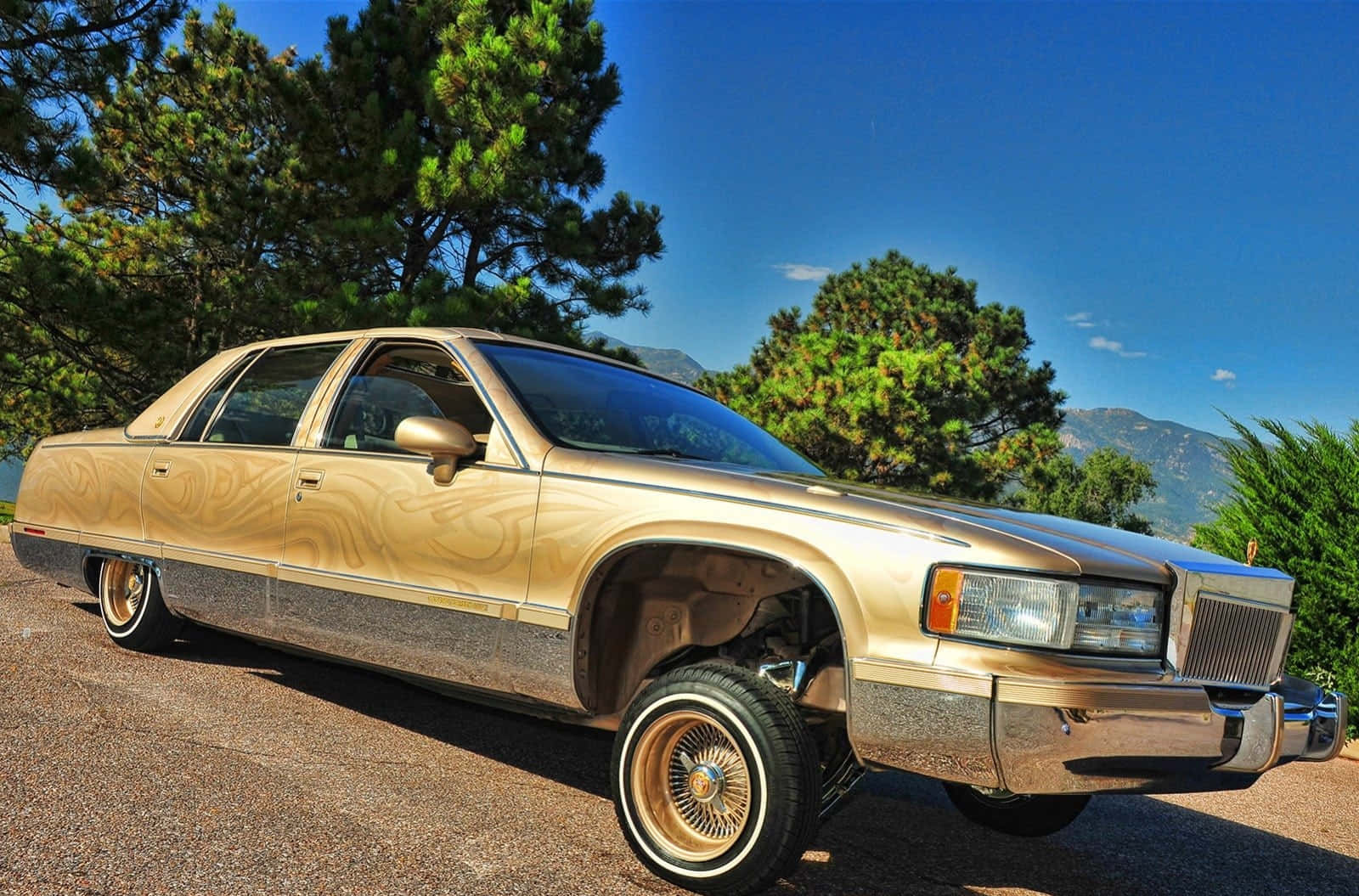 Cadillac Fleetwood 桌布 壁紙 — 一輛金色經典汽車，帶有木紋側板和金色輪圈，停在碎石上，背景是晴朗的藍天、松樹和山脈。