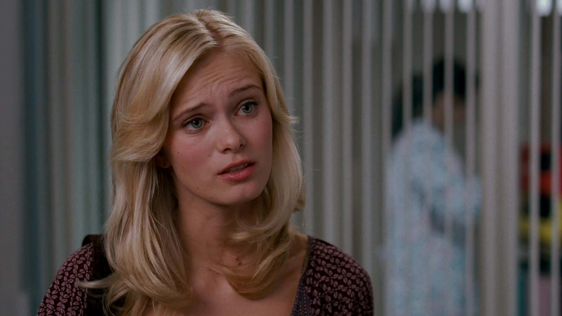 Sara Paxton 桌布 壁紙 — 一位金髮女子帶著憂慮的表情望向鏡頭外，身處一個光線柔和、有垂直百葉簾的室內場景。