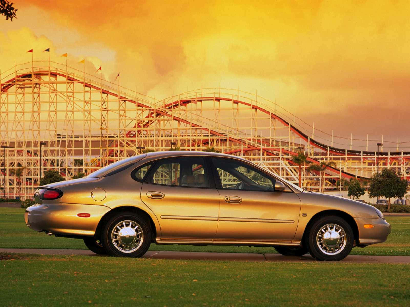 Ford Taurus 桌布 壁紙 — 一輛金色轎車停在草地上，高聳的過山車在火紅的日落天空下閃耀。