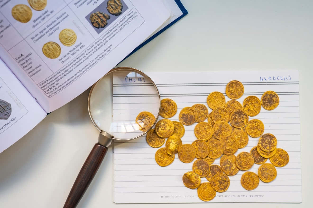 聰明投資，分散風險 壁紙 — 金色古錢幣散落在間條紙上，旁邊有放大鏡和一本打開的錢幣學書籍。