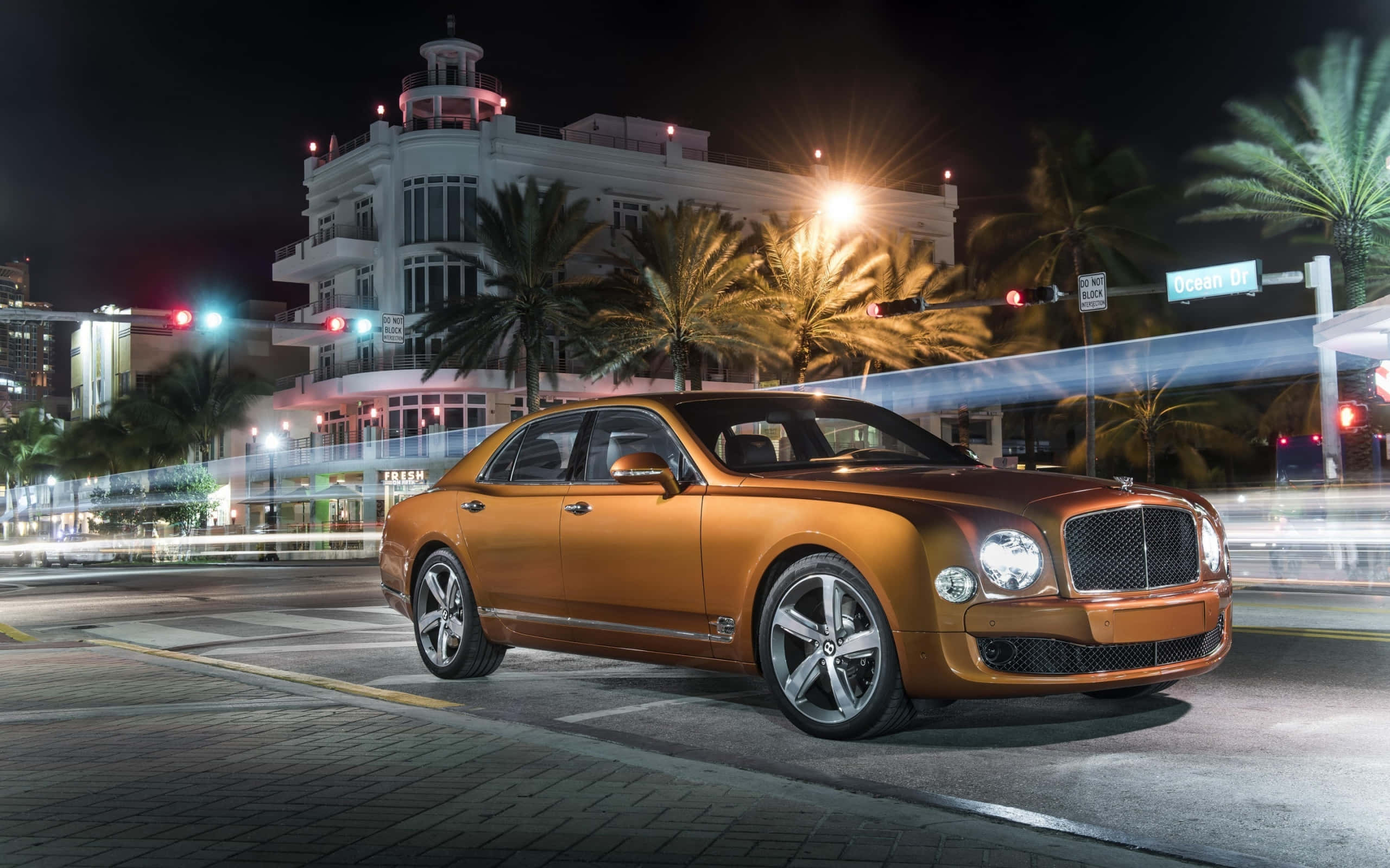 Bentley Mulsanne 桌布 壁紙 — 一輛金色賓利房車在邁阿密熱鬧的夜晚街道上滑行，街道兩旁種滿棕櫚樹和閃閃發光的裝飾藝術建築。