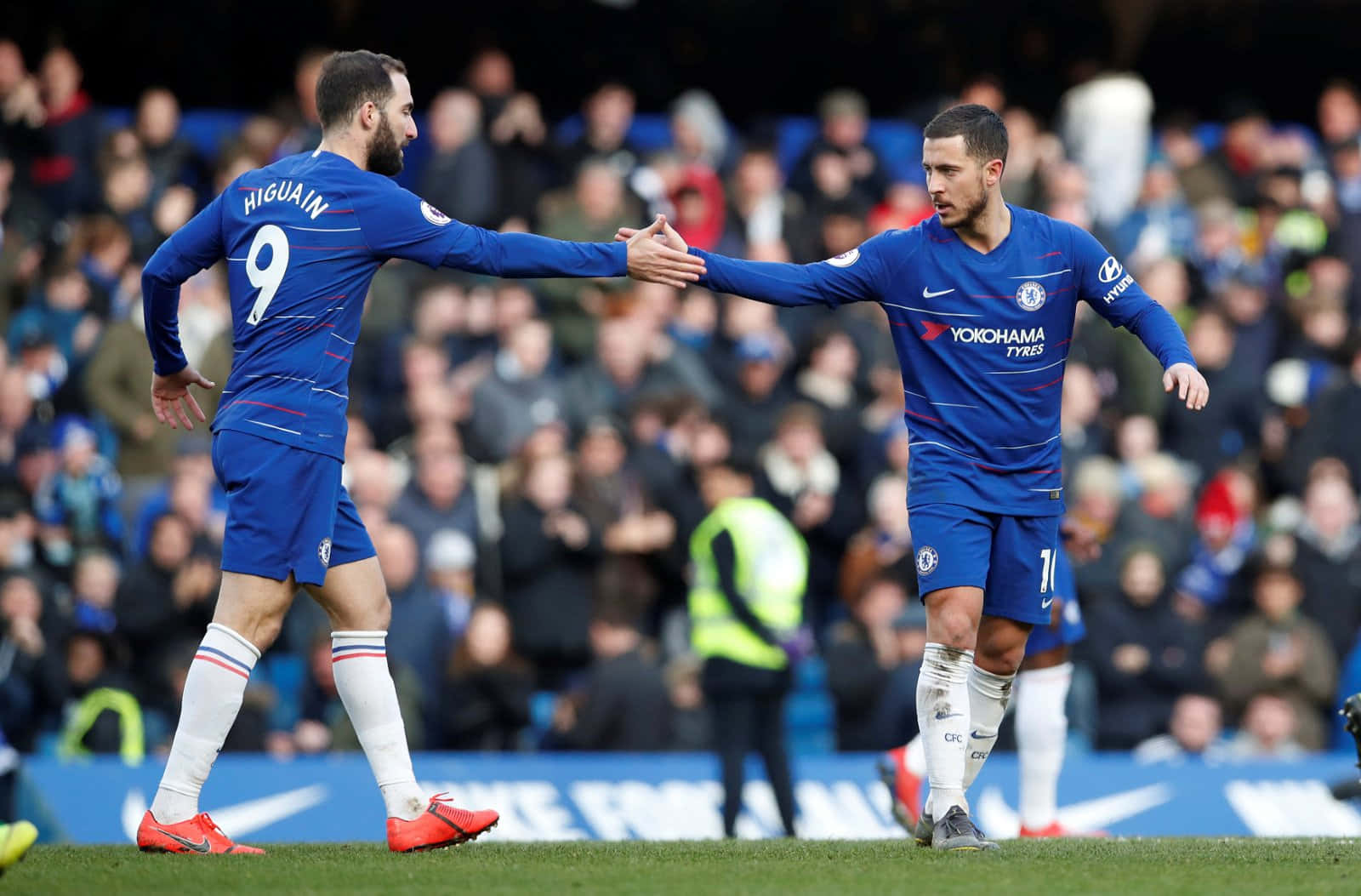 Gonzalo Higuain 與Eden Hazard 握手 壁紙 — 兩名身穿藍色球衣的車路士足球員在草地上握手，背景有模糊的觀眾。