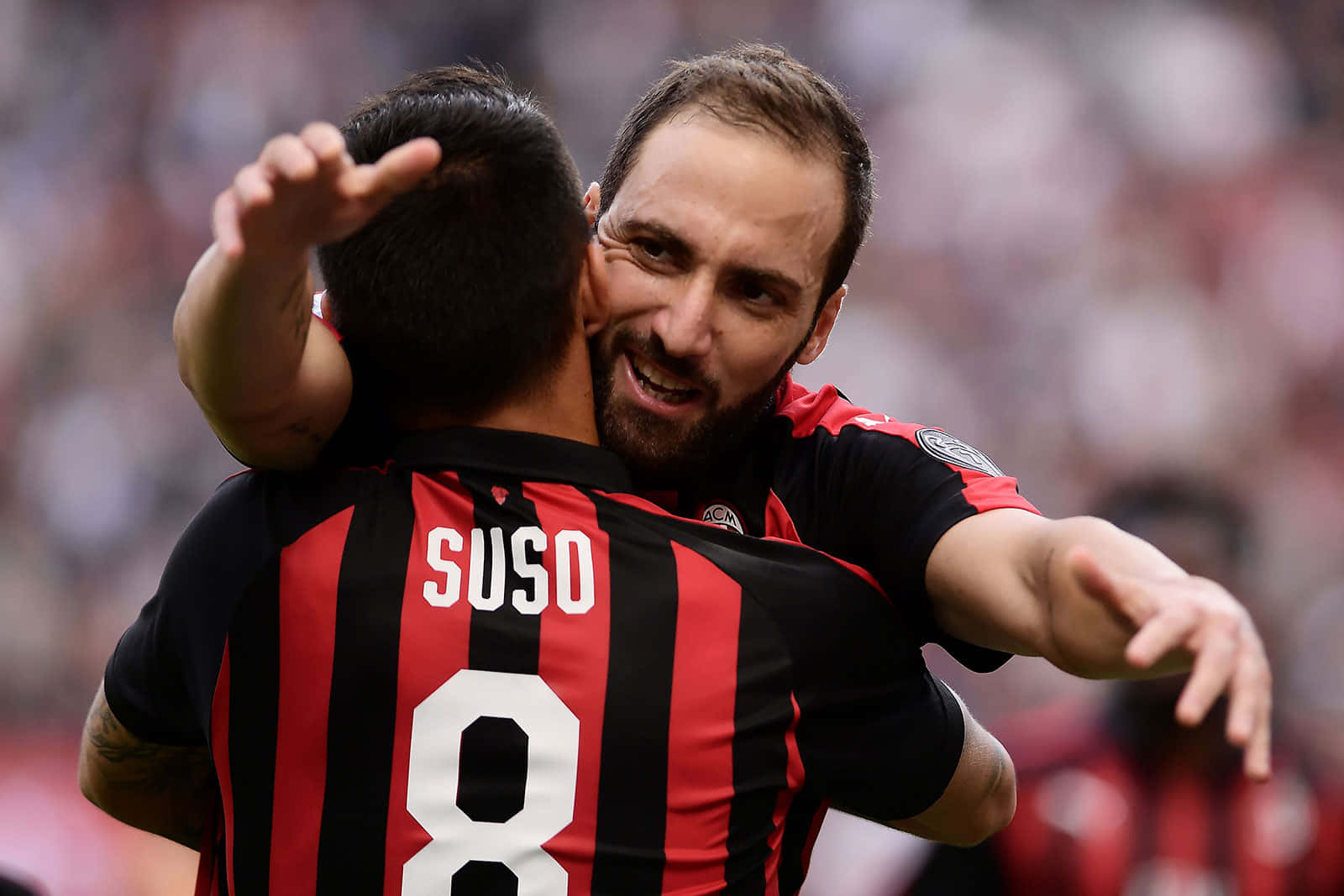 Gonzalo Higuain擁抱隊友Fernandez Suso 壁紙 — 兩名身穿紅黑間條球衣的足球運動員在球場上開心地擁抱。