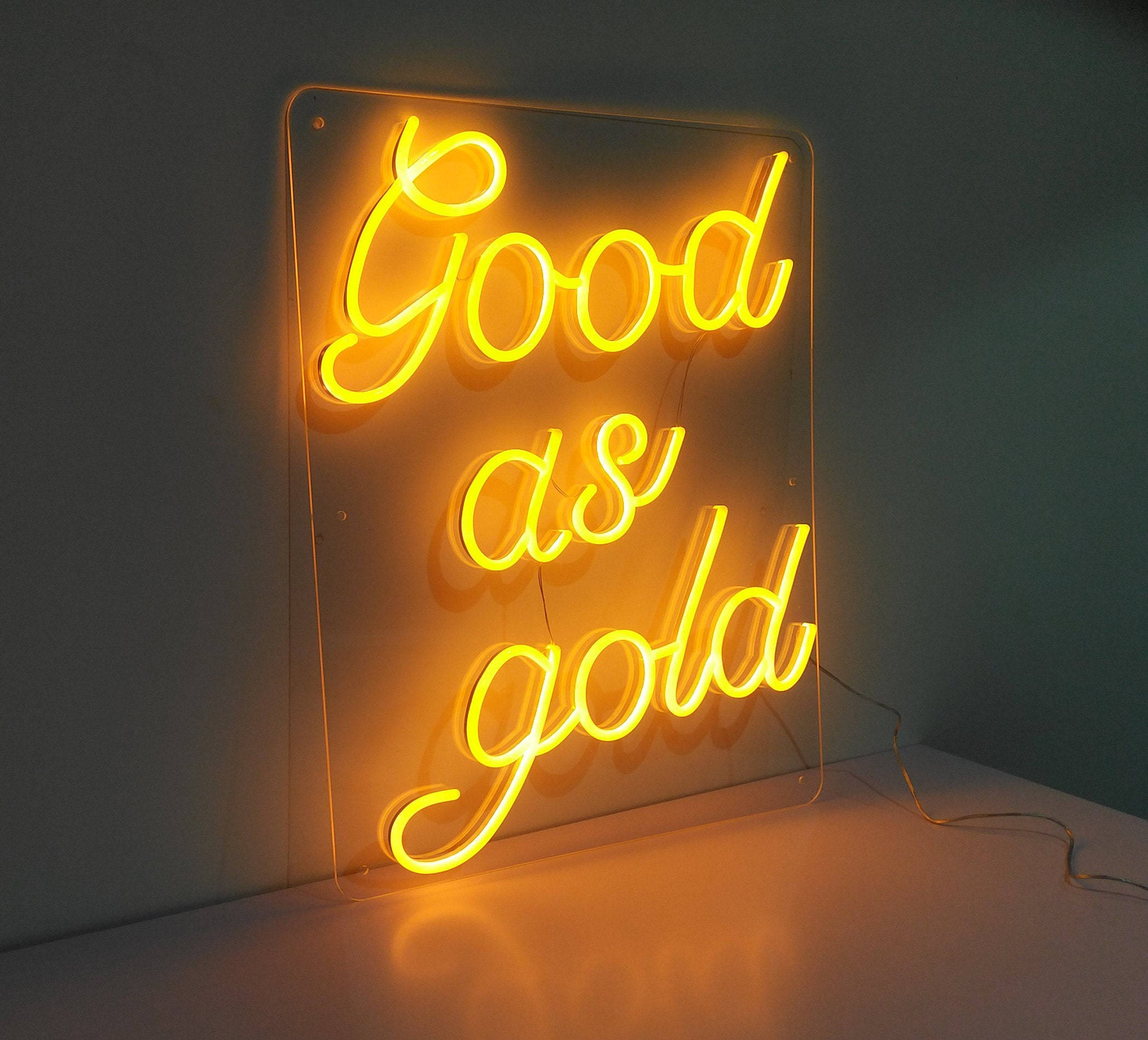 「Good As Gold」霓虹黃金語錄 壁紙 — 一個發光的黃色霓虹燈牌，上面寫著「Good as gold」，在樸素的牆壁和表面上投射出溫暖的光芒。